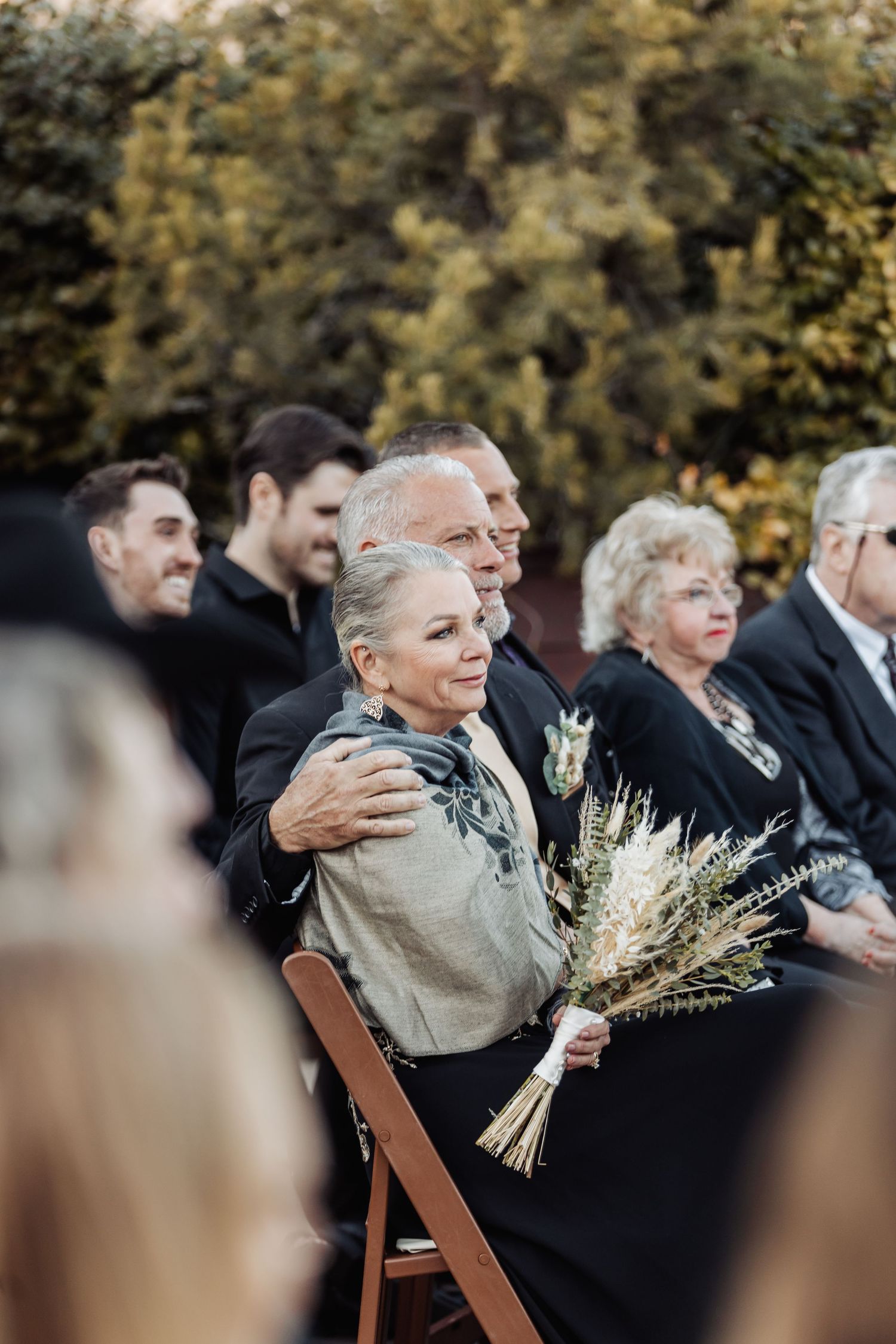 Wedding guests seated in wooden chairs at an outdoor ceremony with autumn foliage in the background.