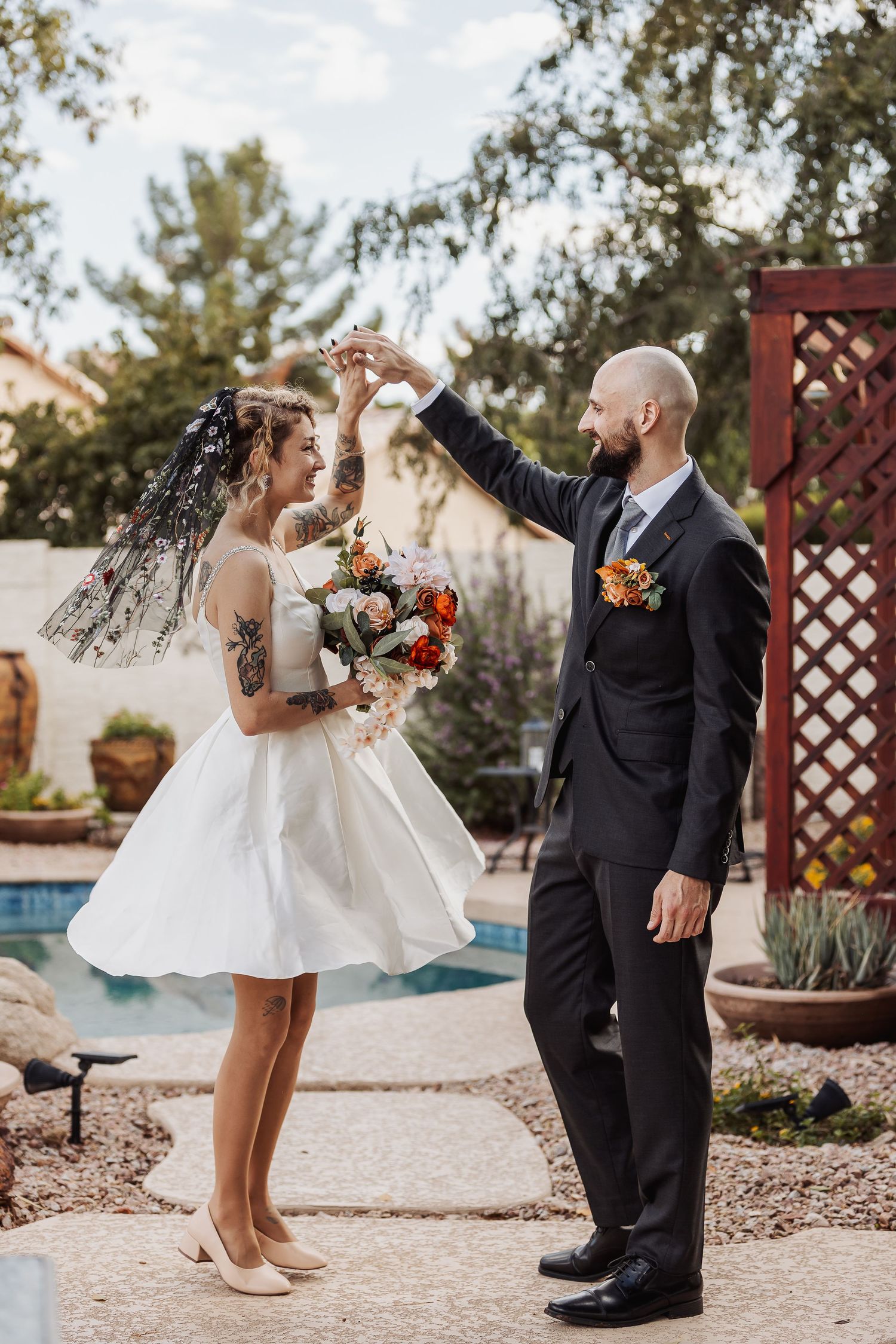 Wedding couple poses in a garden setting with decorative wooden lattice and pool in the background.