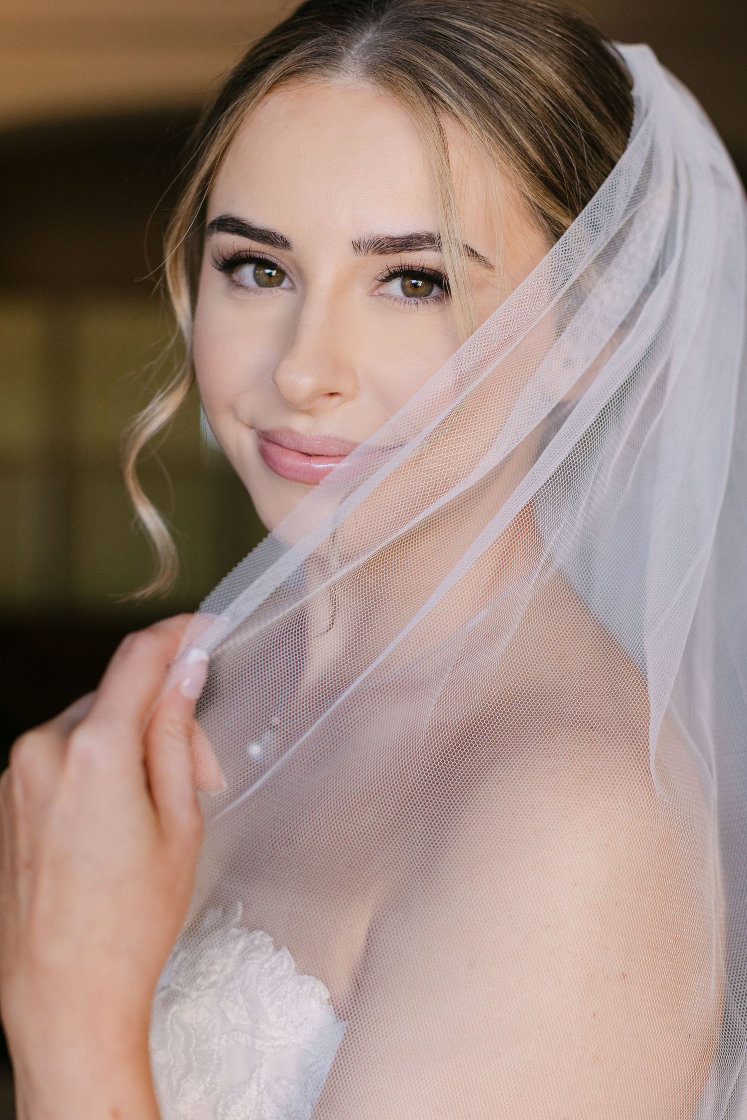 A bride wearing a delicate white veil poses for romantic wedding portraits in soft lighting.
