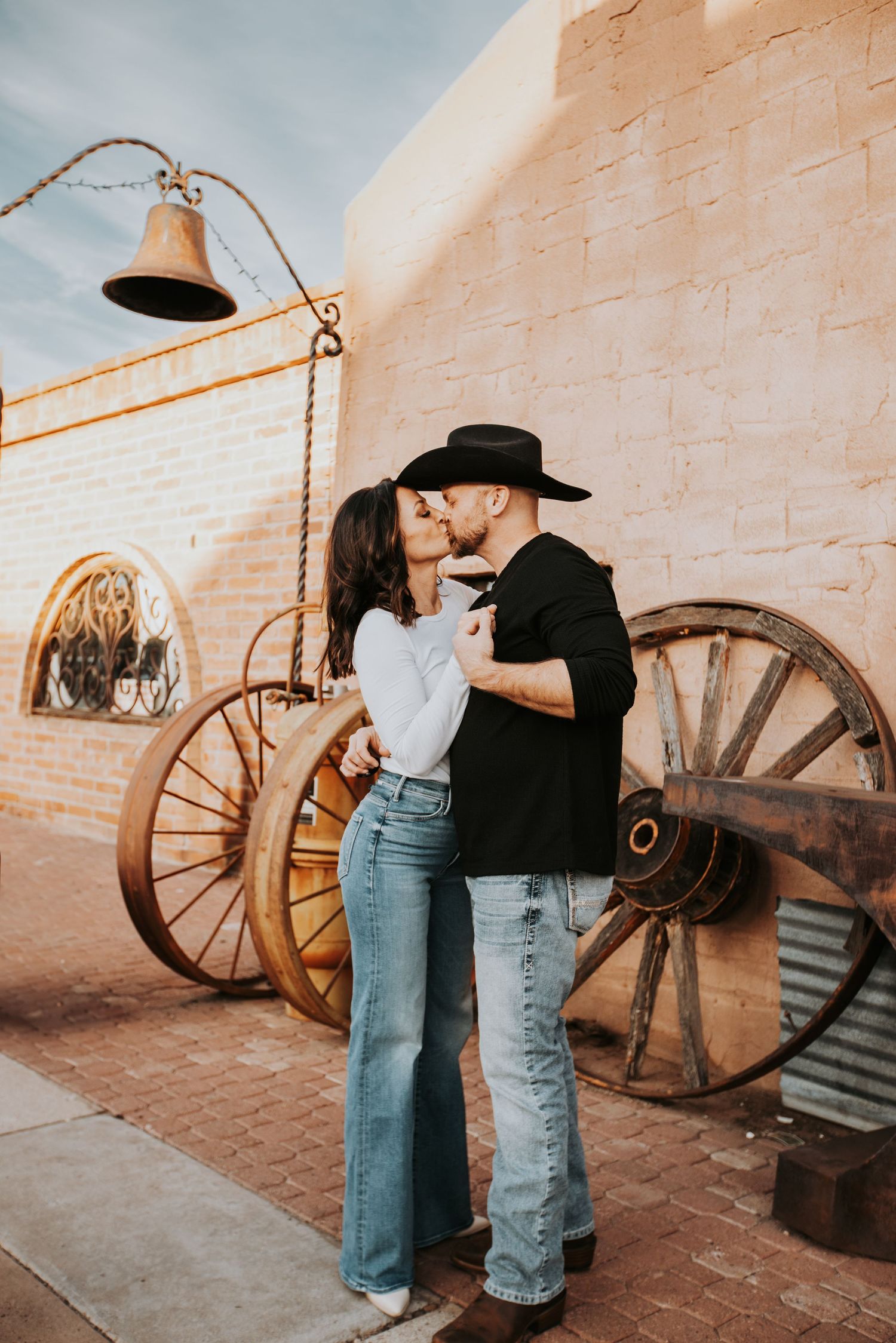 Romantic western themed couple embracing in front of rustic brick wall with vintage wagon wheels and lantern.