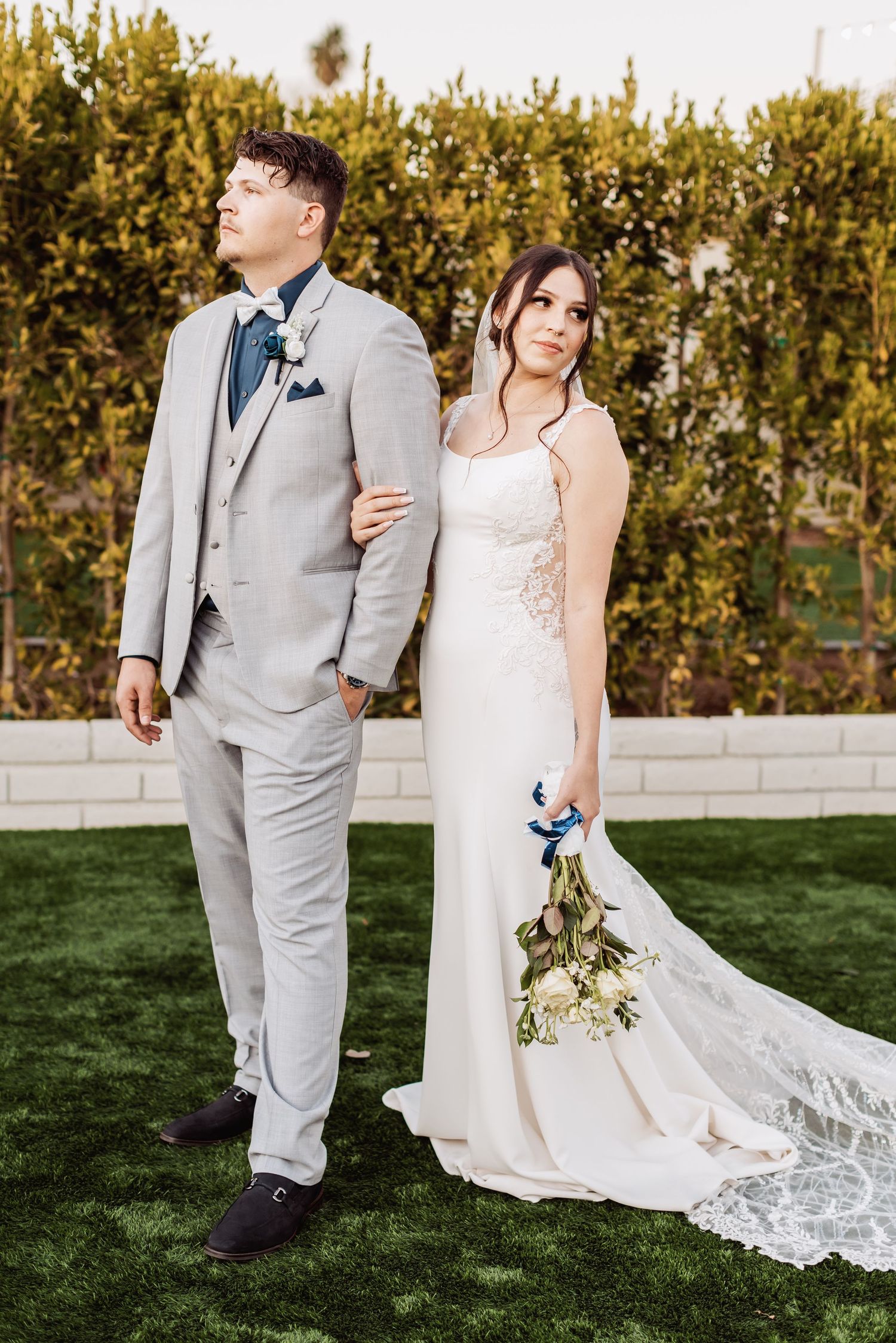 Elegant wedding couple walking outdoors in formal attire with a flowing white dress train on green grass.