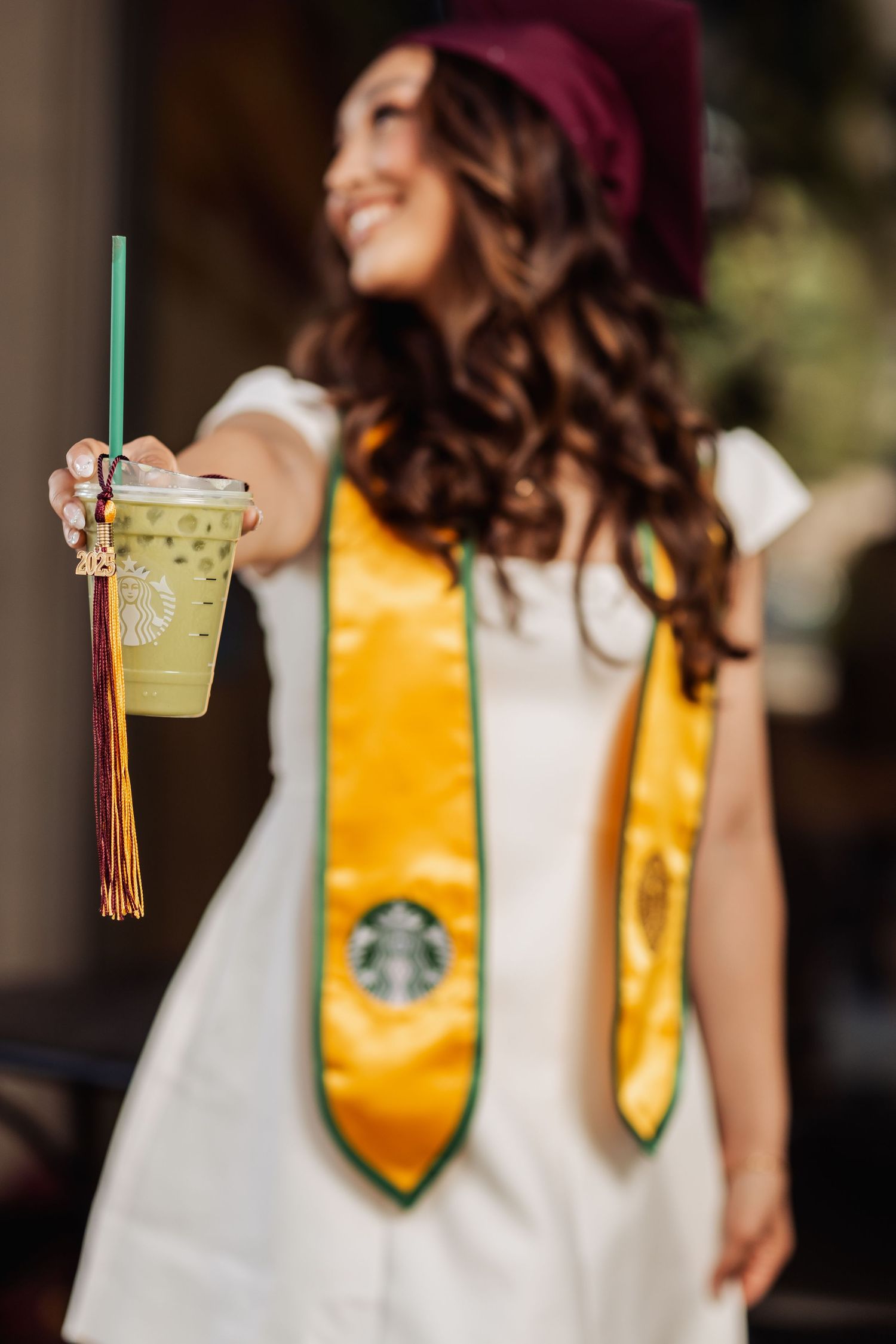 Graduate holds Starbucks drink while wearing white dress, yellow honors stole, and burgundy graduation cap outdoors.