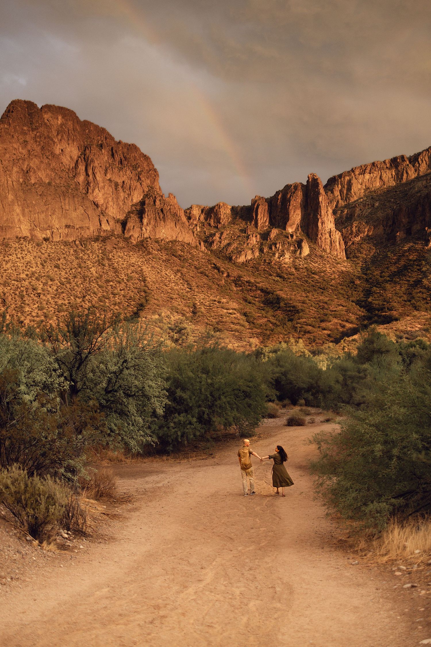 Two hikers walk down a desert trail at sunset with towering cliffs and a rainbow in the Superstition Mountains of Arizona.