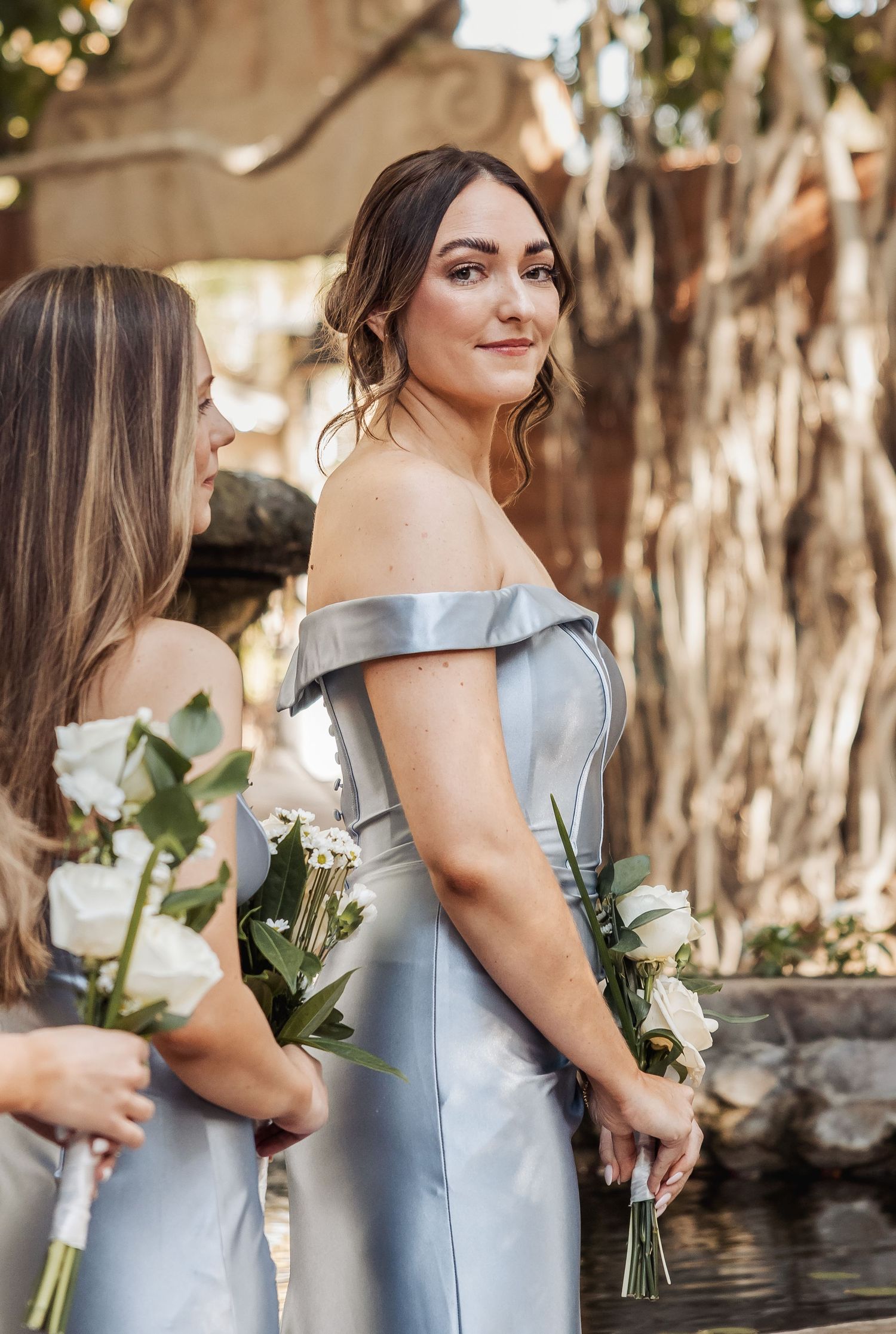 Bridesmaid in off-shoulder silver satin dress holds white roses at outdoor wedding venue with natural lighting.