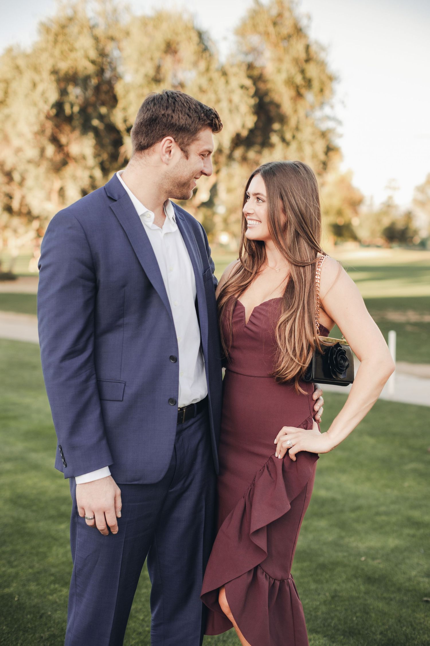 A couple shares a romantic moment outdoors, with her in a burgundy dress and him in a navy suit against autumn trees.