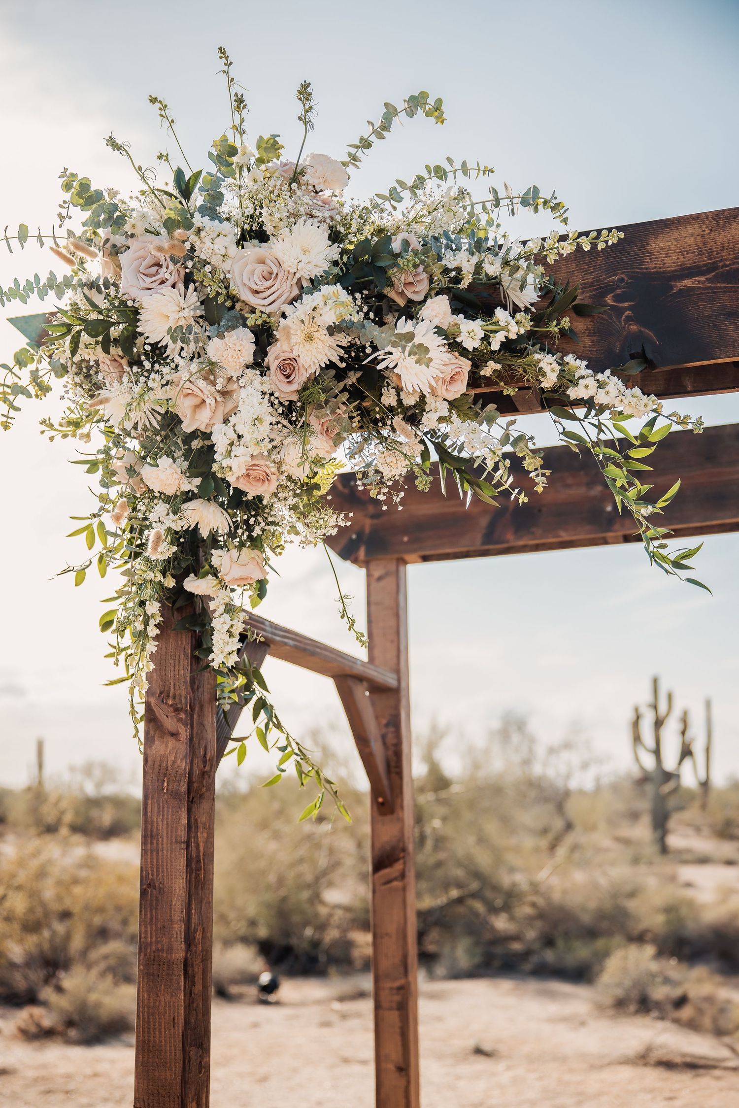 Rustic wooden wedding arch decorated with white and blush roses against a desert landscape with cacti in the background.