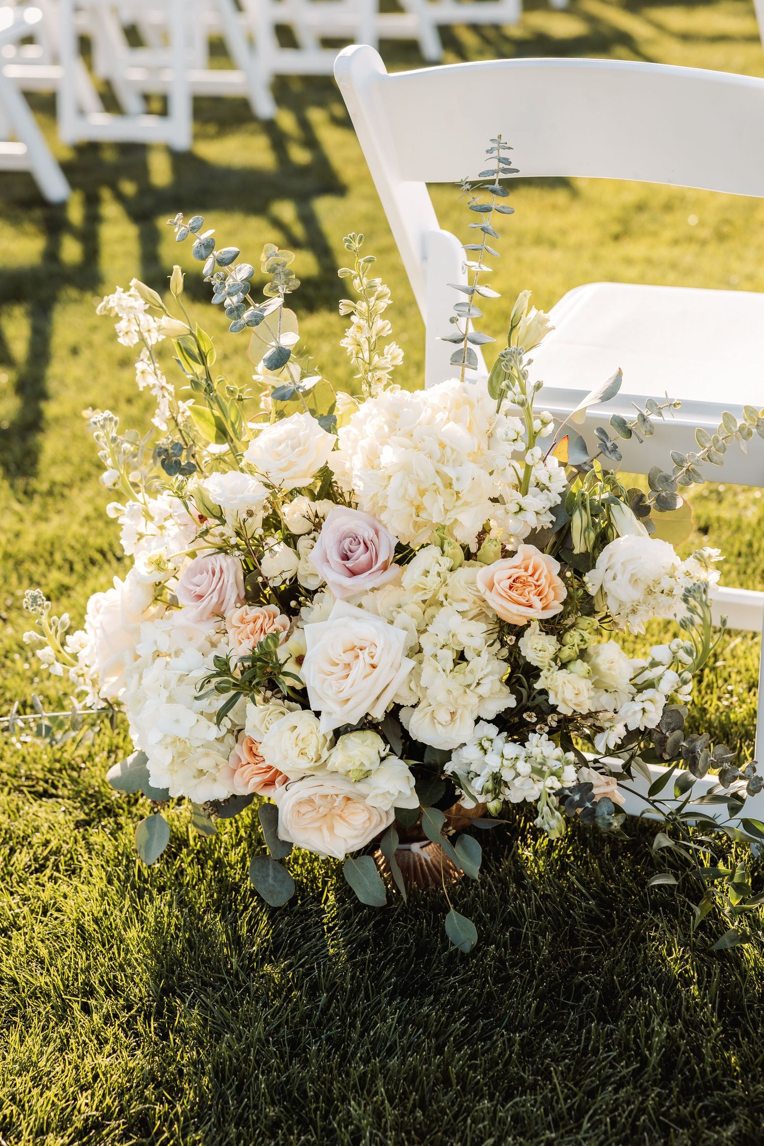White and blush florals with eucalyptus rest on lush grass in golden sunlight at an outdoor event.