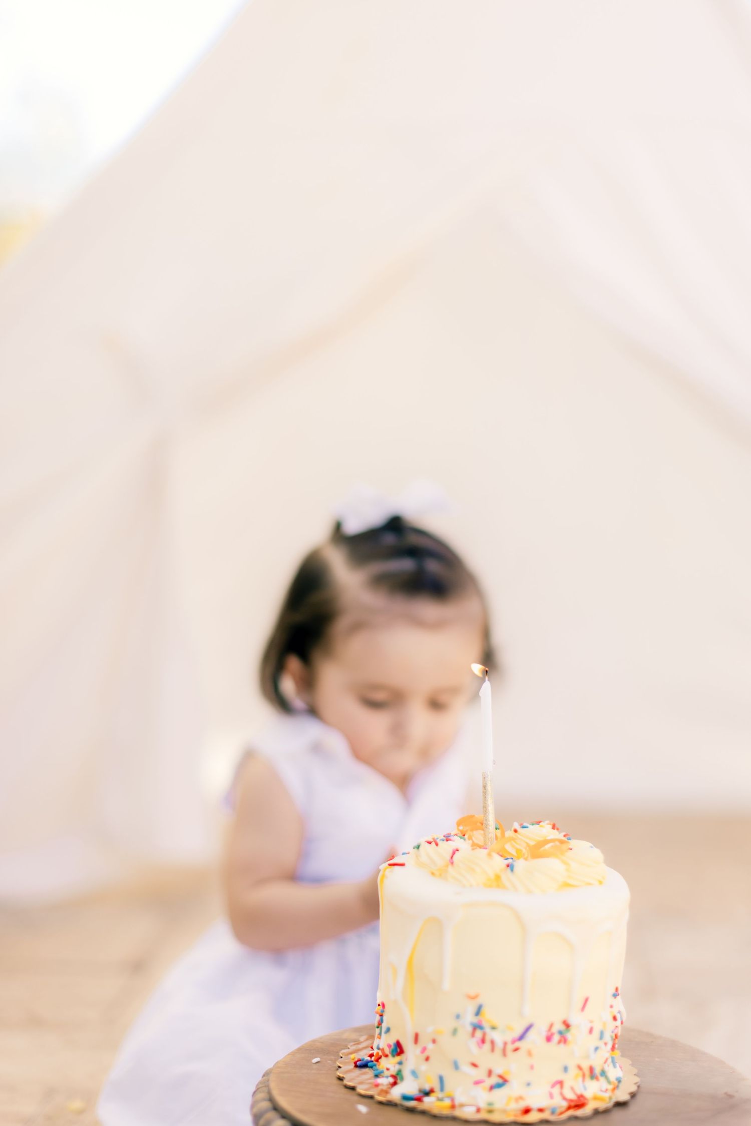 A white birthday cake with sprinkles and decorations sits on a wooden surface near a blurred background.
