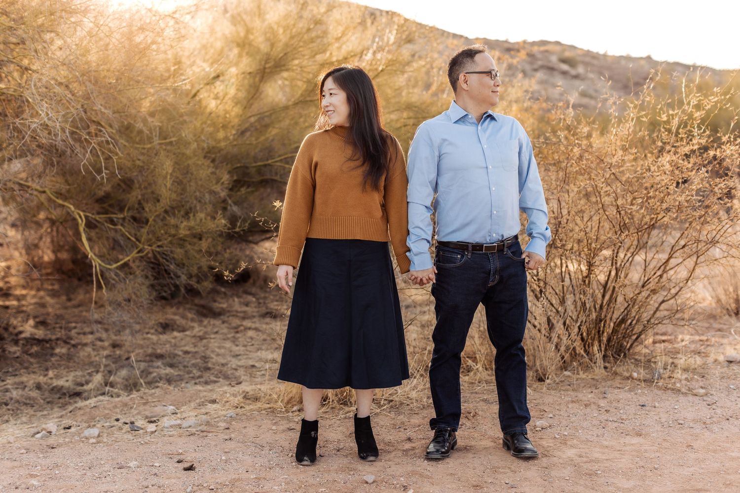 Two people in casual attire walking together through a desert landscape with golden brush and vegetation at sunset.