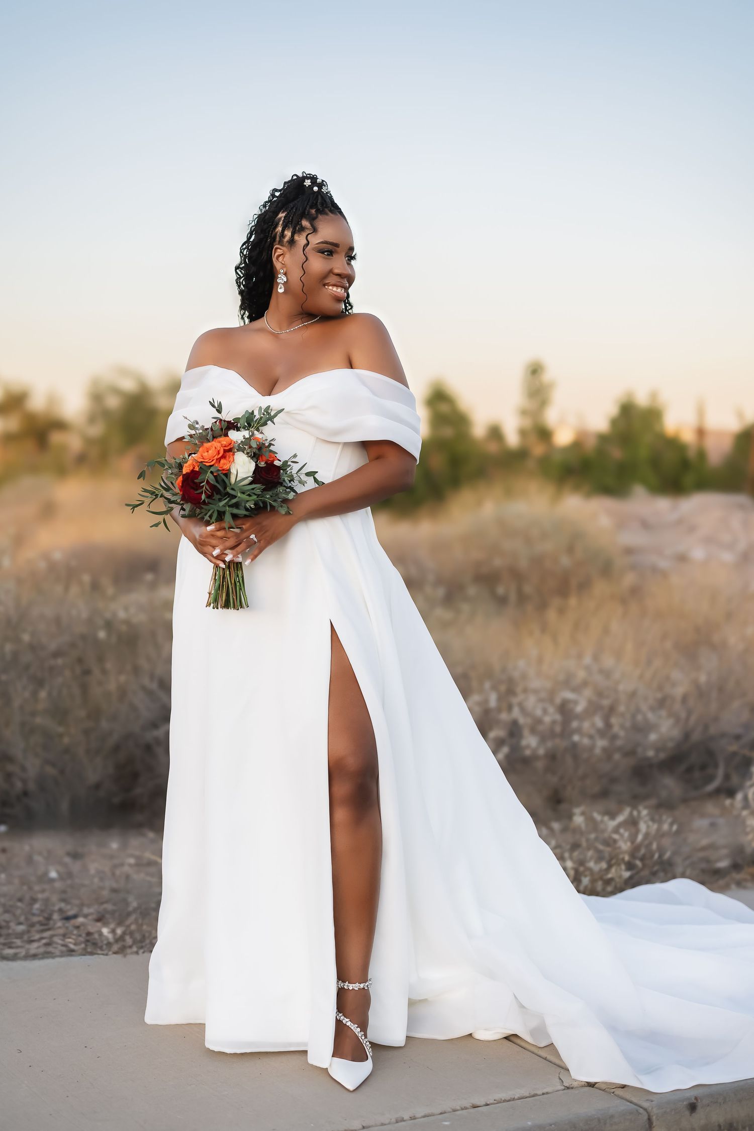 Elegant bridal portraits in a desert setting at sunset with flowing white off-shoulder gown and floral bouquet.