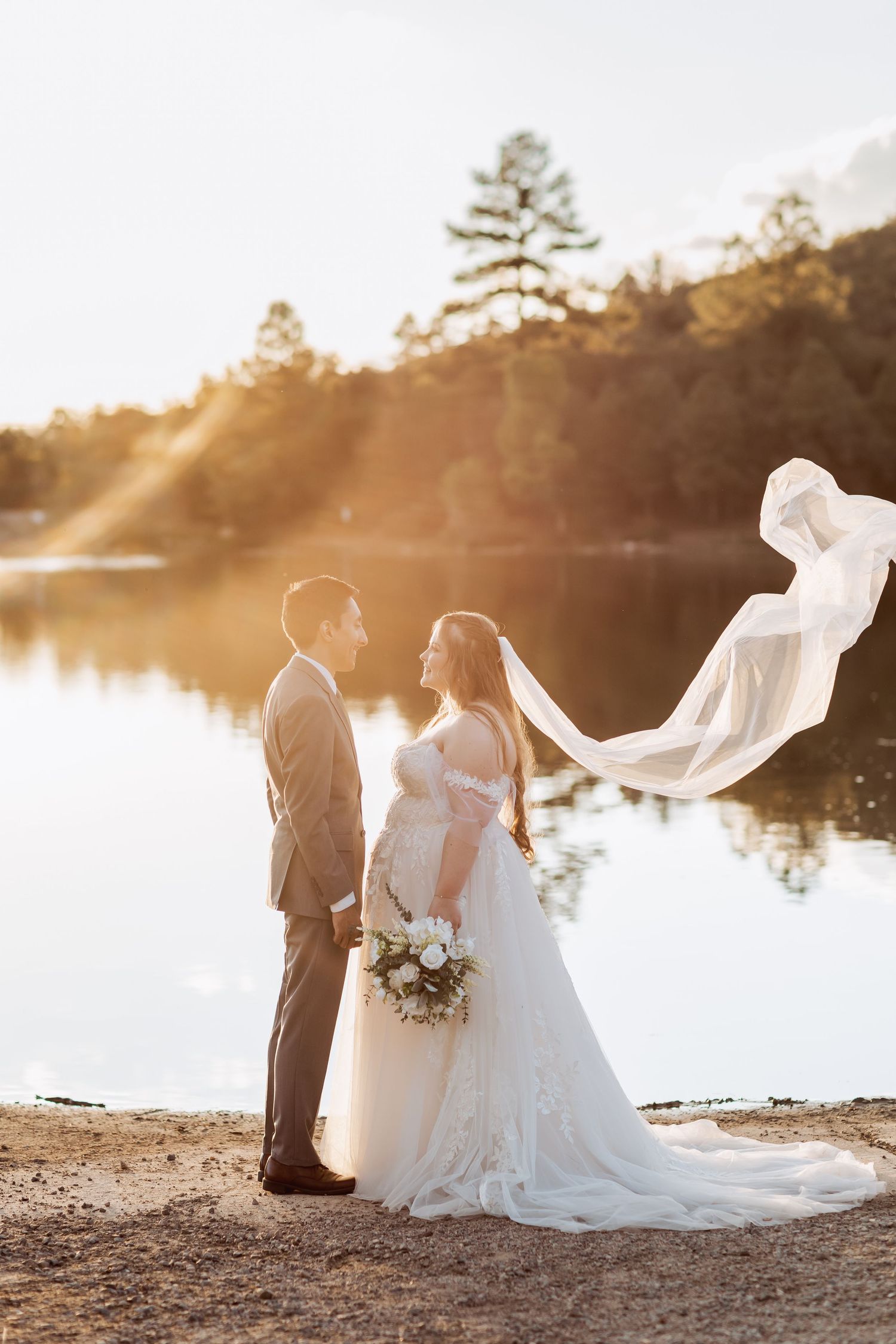 Romantic sunset wedding photos of couple by a lake with flowing veil in golden light.
