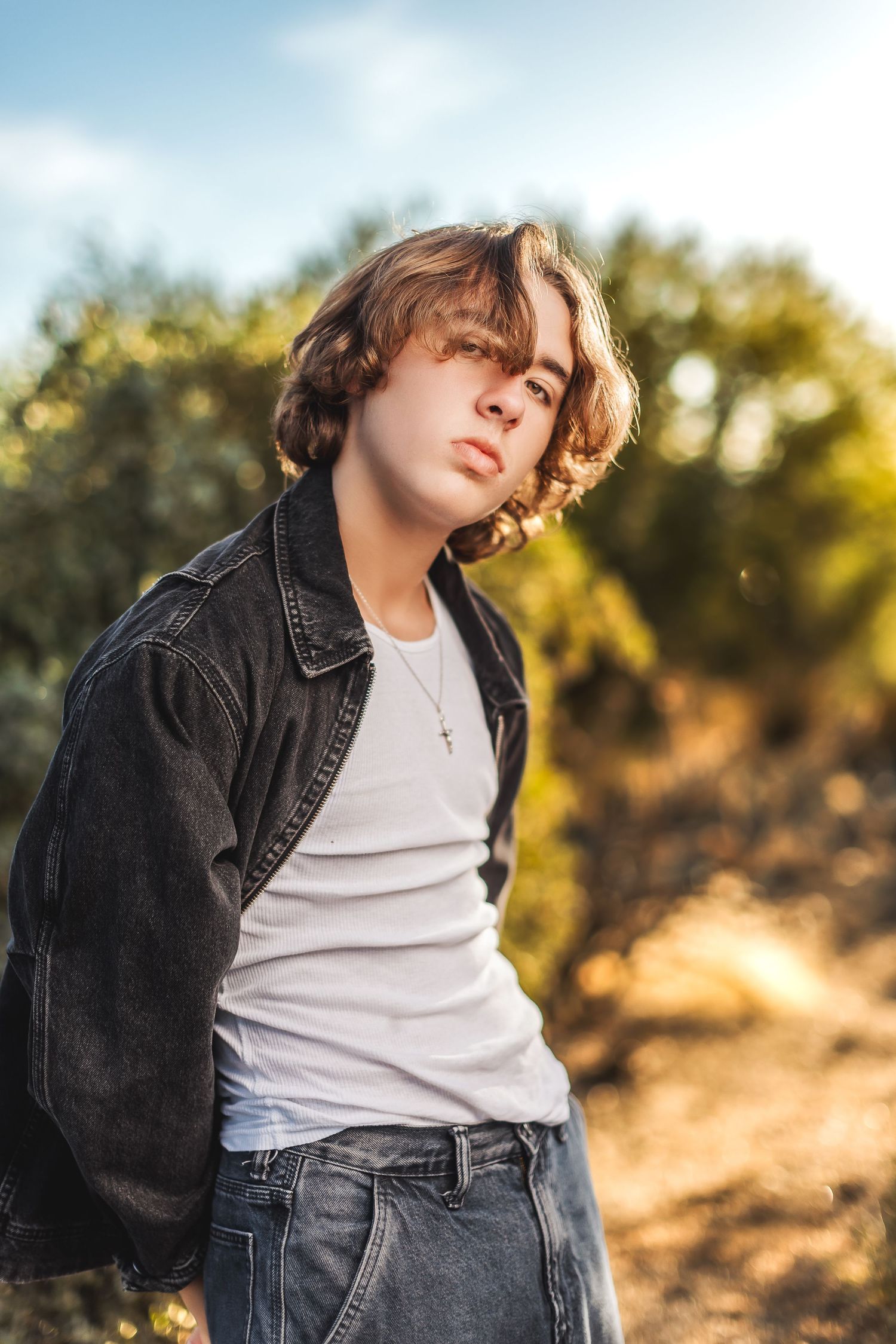 Fashion portrait of person in denim jacket and white tank top posing outdoors against natural sunlit background.