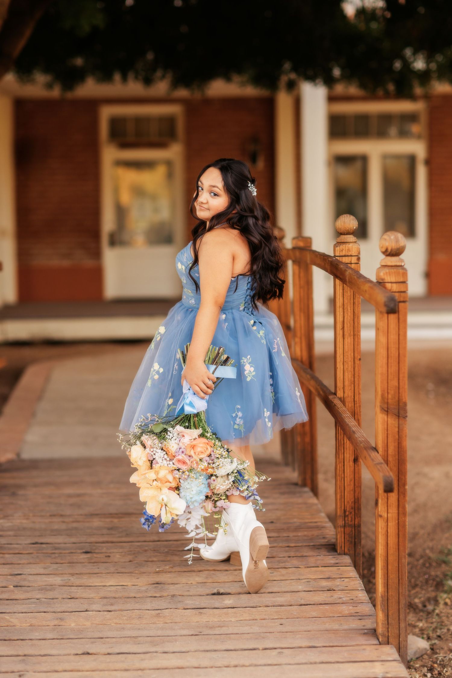 A woman in a blue floral dress poses on a wooden bridge while holding a pastel bouquet of flowers.