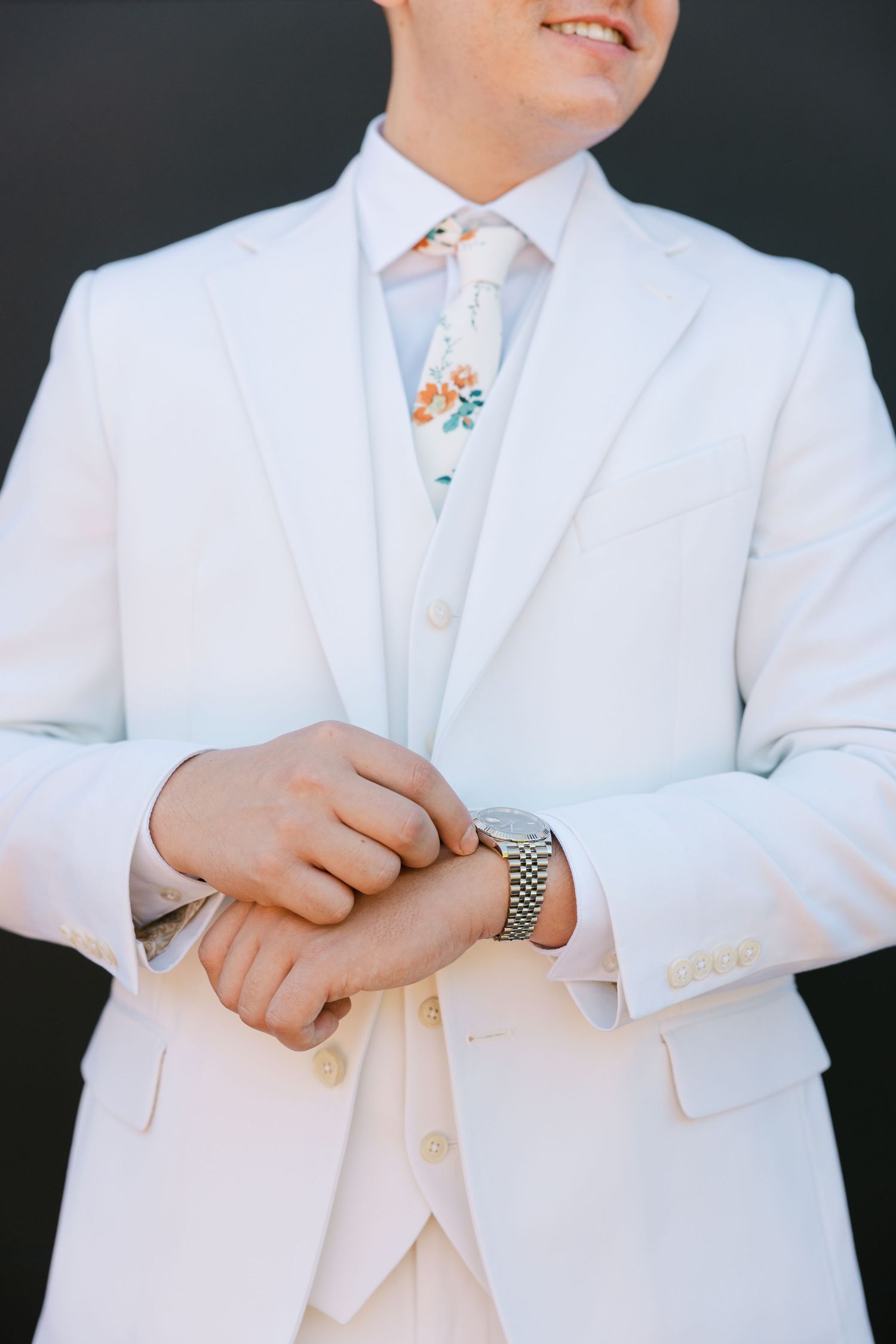 Close-up details of a white formal suit showing lapels, buttons, and a wristwatch accessory against a dark background.