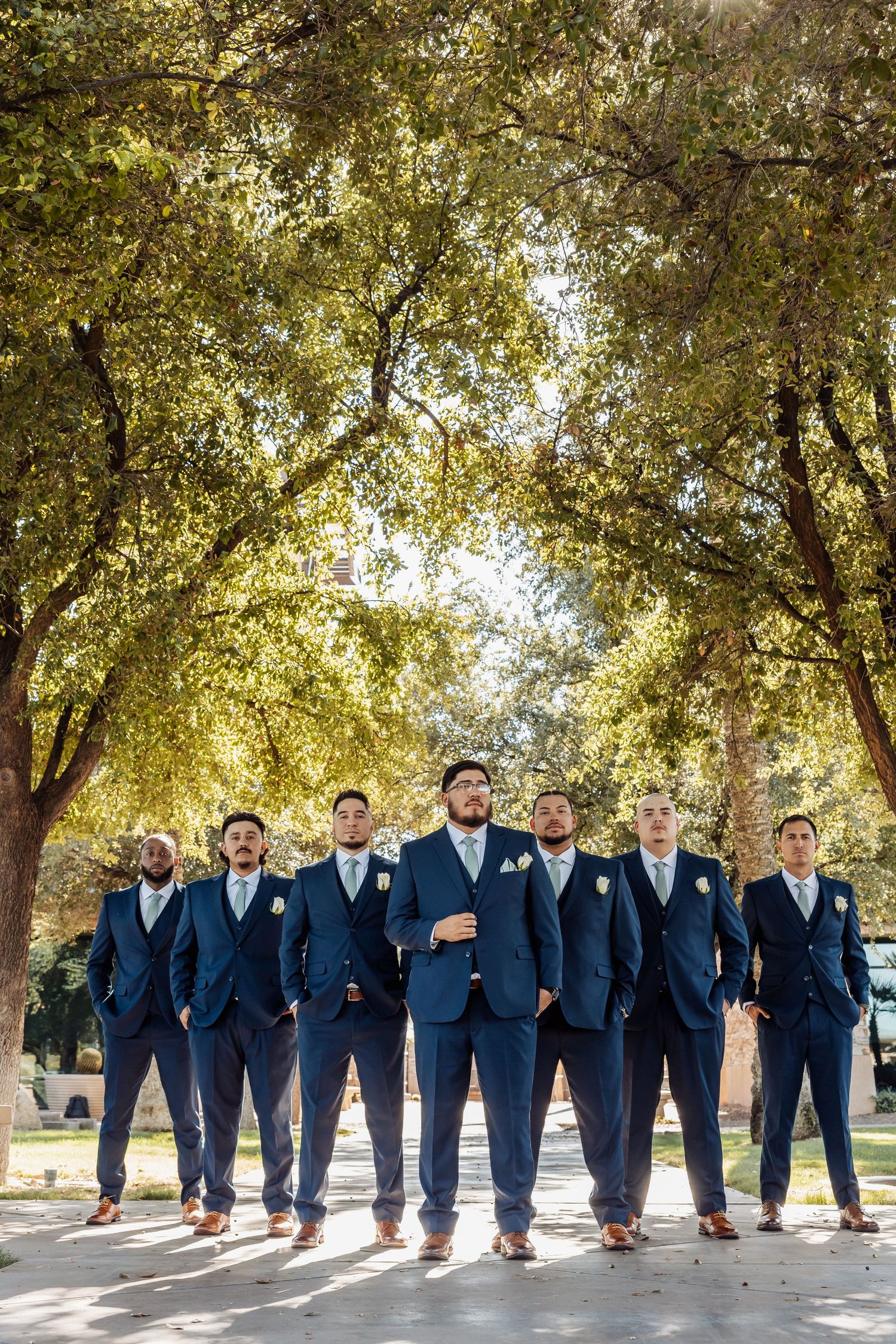 Groomsmen in navy blue suits walking and posing under tree canopy in outdoor setting.