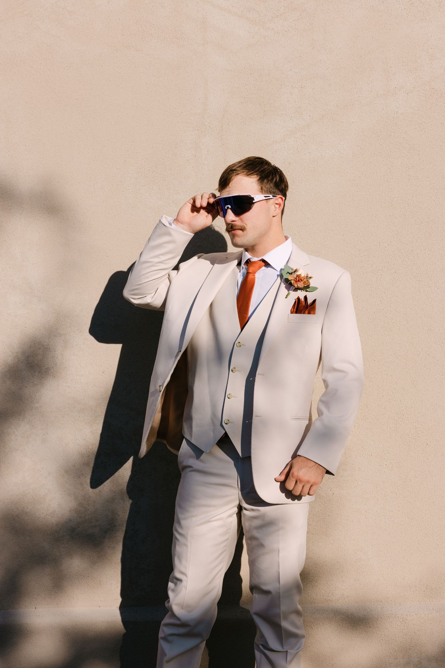 A person in a light beige suit with orange tie and sunglasses poses against a wall casting dramatic shadows.