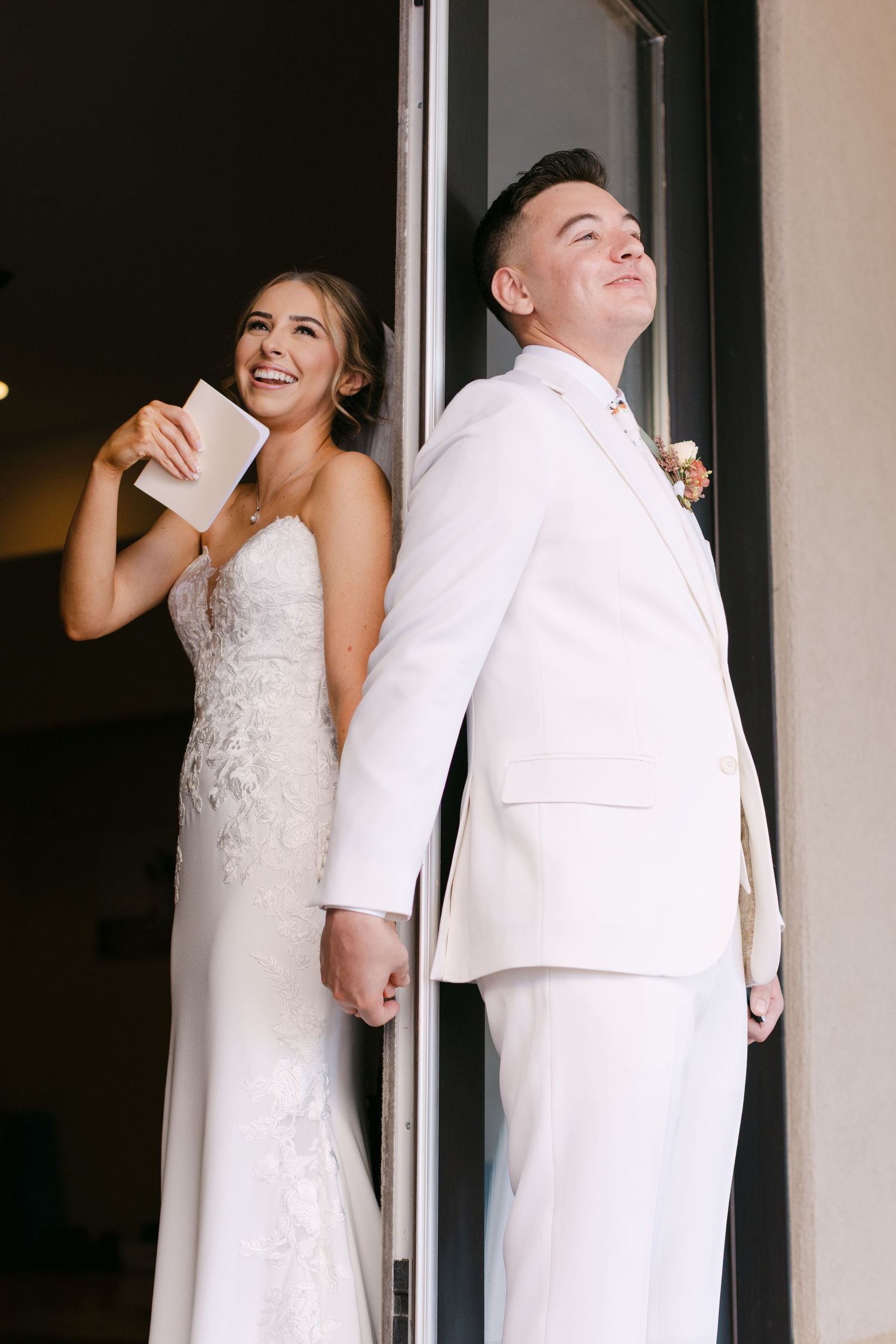 A couple in wedding attire stands back to back in a playful first look moment before their ceremony.