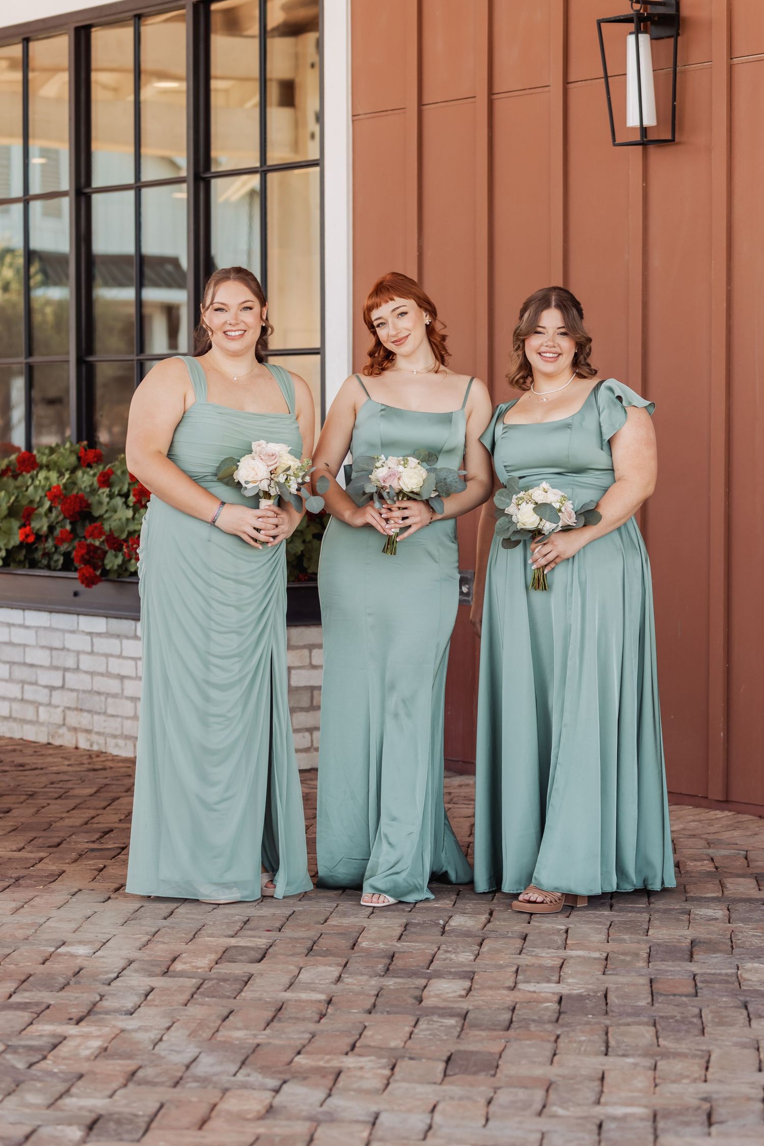 Bridesmaids in matching sage green dresses holding bouquets pose together on brick pavement.