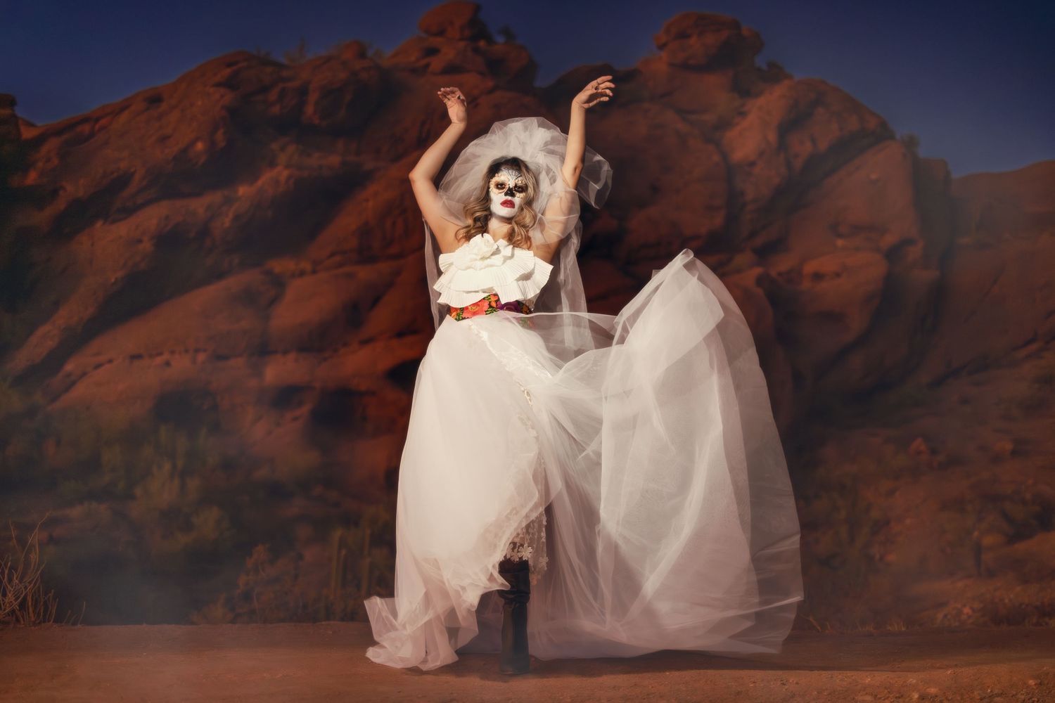 Figure in flowing white dress poses gracefully against dramatic red rock formations in desert landscape at sunset.