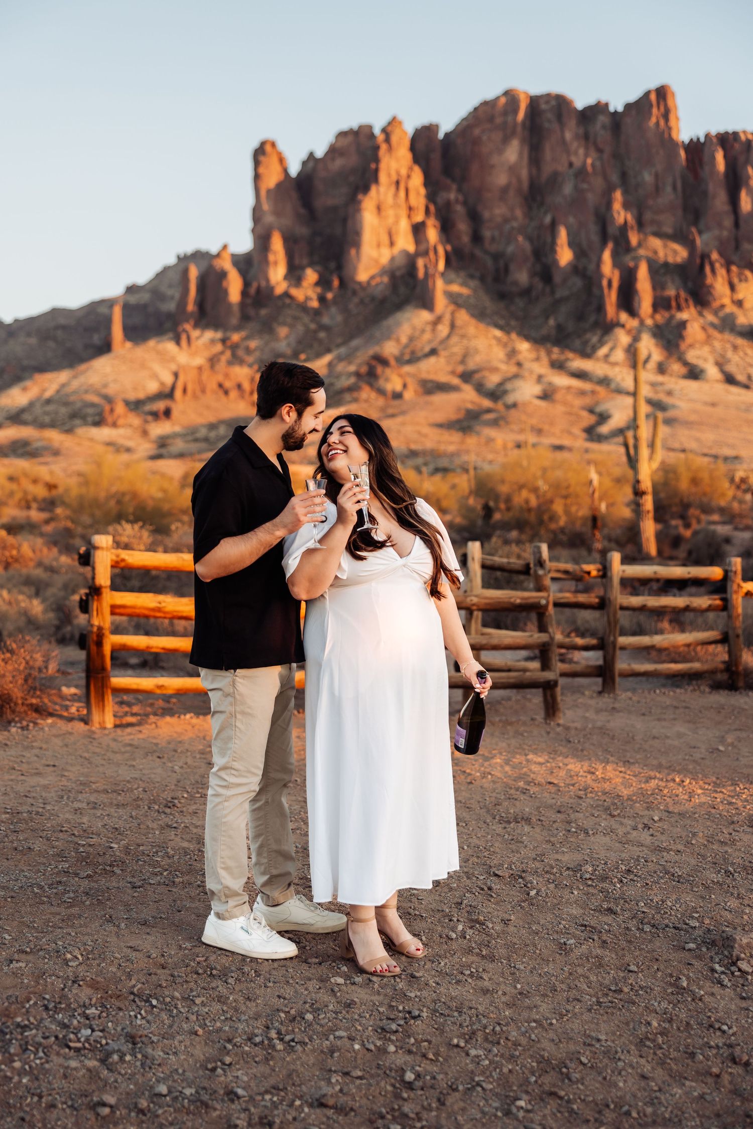 Couple shares intimate moment at sunset near Superstition Mountains in Arizona with wooden fence and desert landscape.