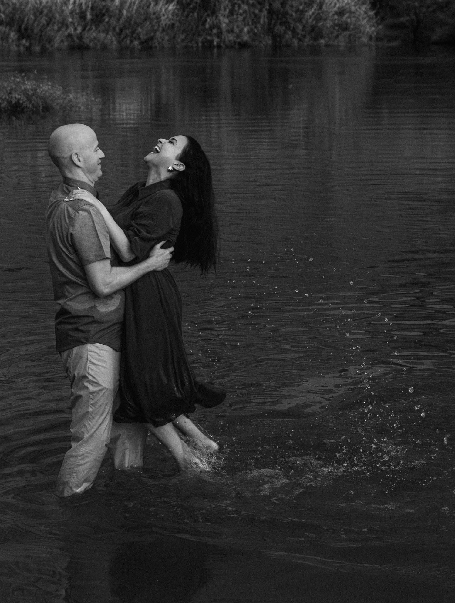 A couple embraces while standing in rippling water, captured in dramatic black and white photography.