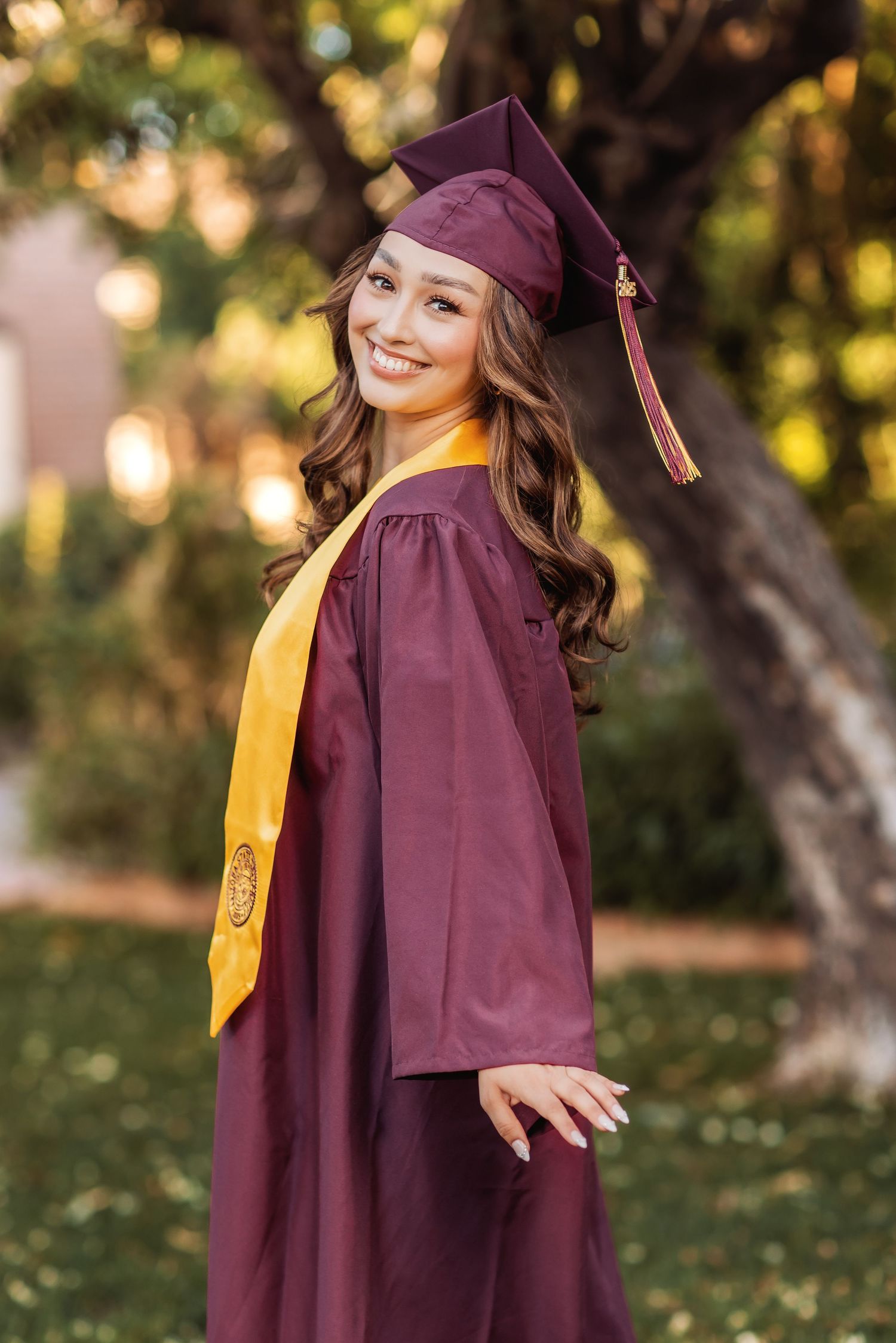 Graduate in burgundy cap and gown poses joyfully outdoors during golden hour graduation photoshoot.