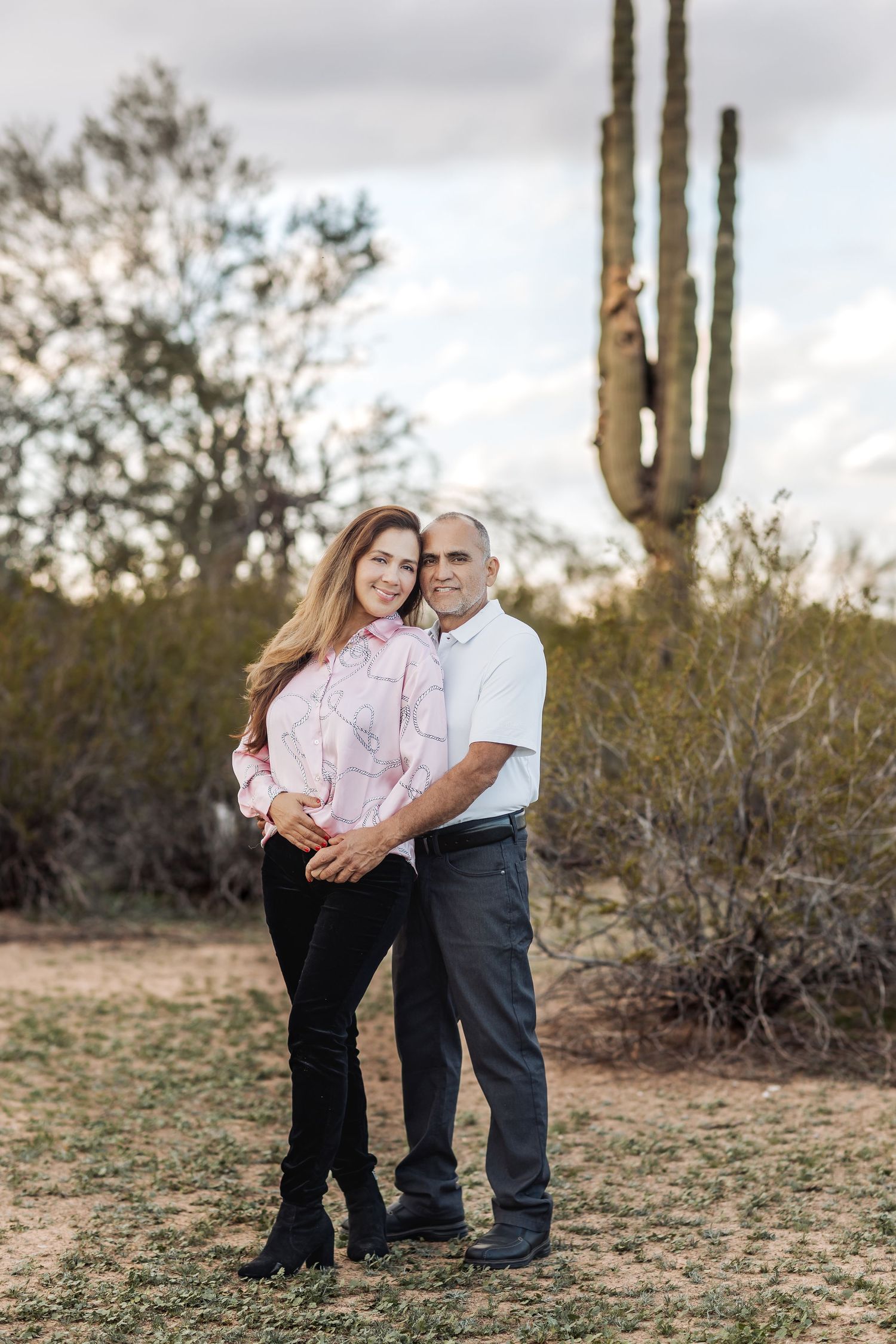 A couple embraces in front of a tall saguaro cactus in the Arizona desert during golden hour.