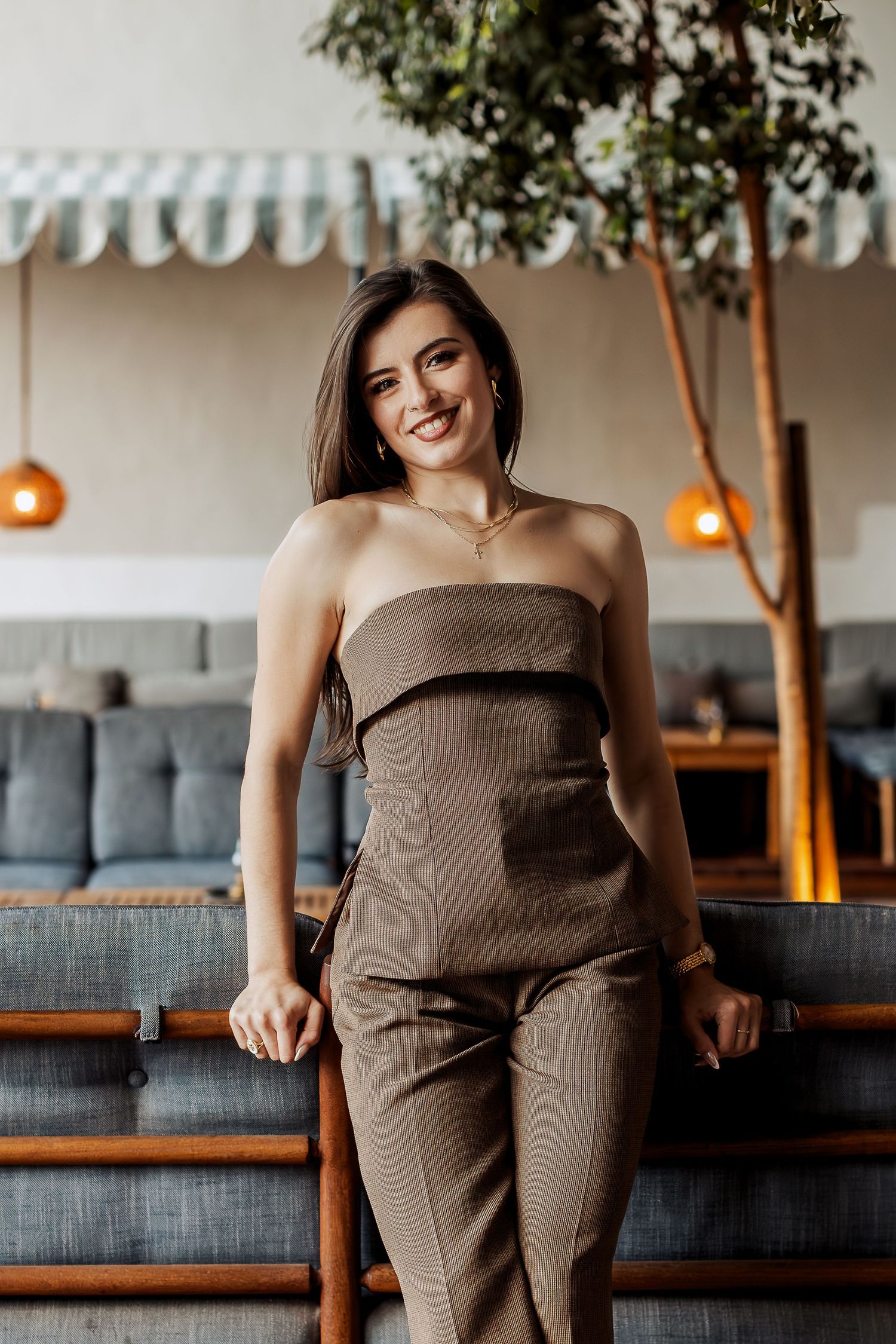 A person in a brown strapless jumpsuit poses in front of a modern restaurant interior with pendant lights and bench seating.