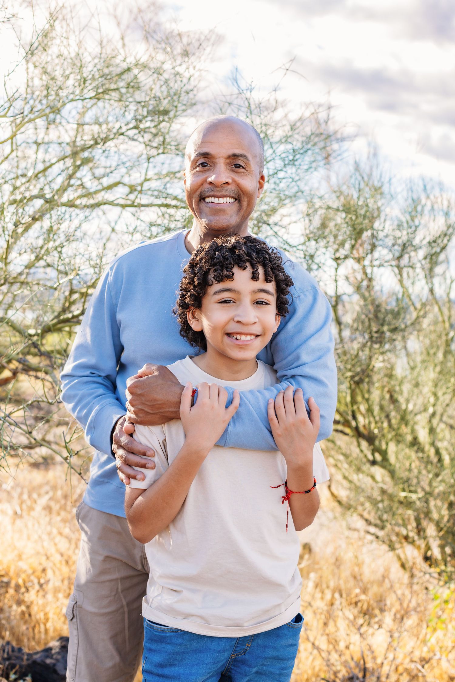Smiling grandparent and grandchild embrace outdoors against backdrop of desert brush and cloudy sky.