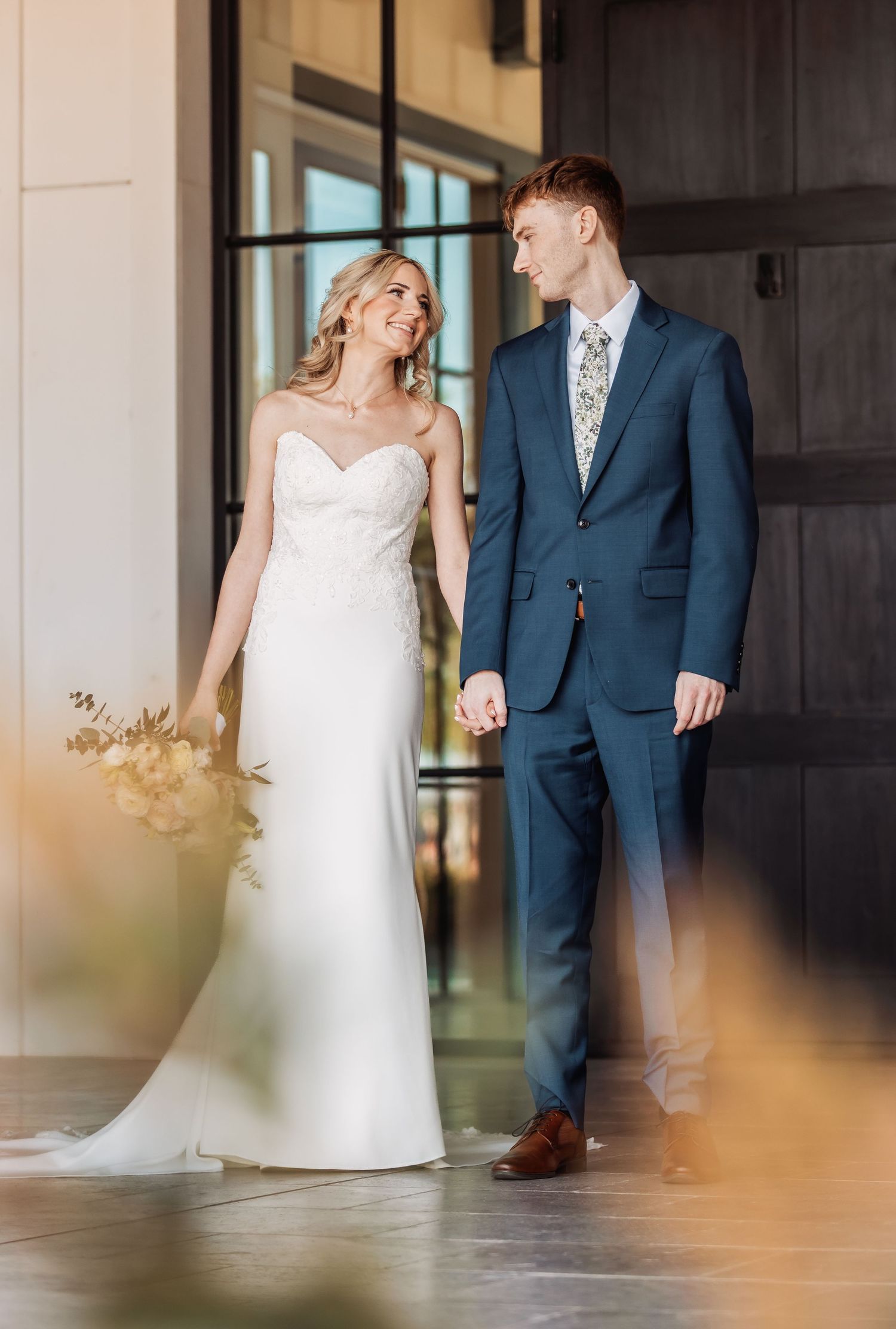 A couple in wedding attire shares a candid moment in a sunlit hallway during their first look.