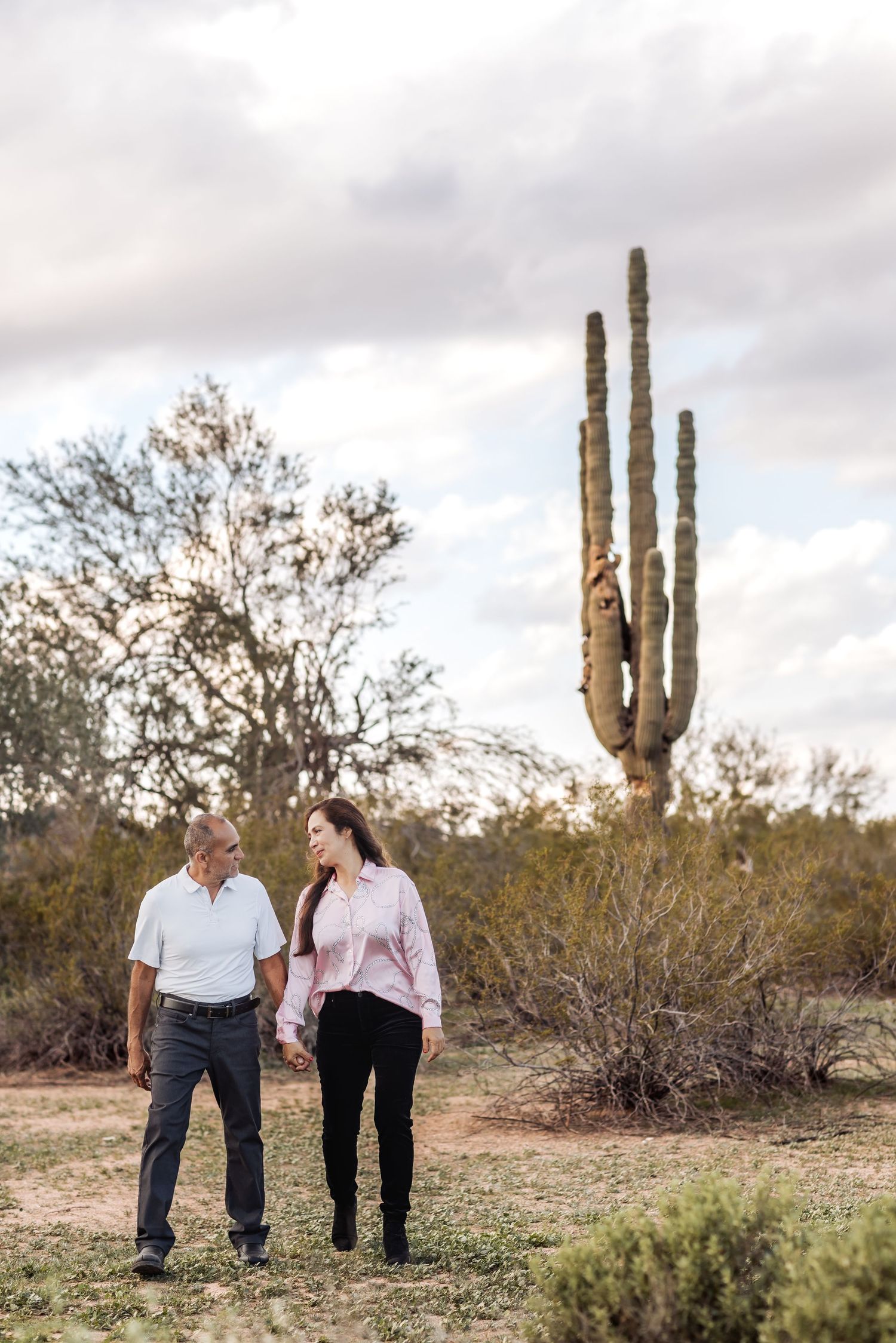 A couple stands together in a desert landscape with tall saguaro cacti and desert brush under cloudy skies.