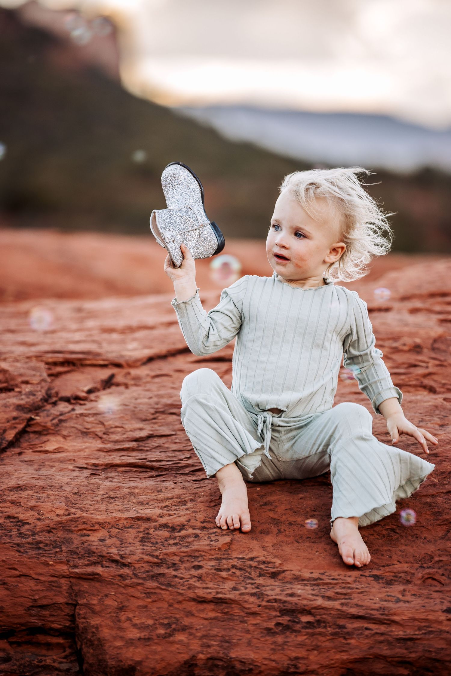 Small child in light gray outfit sits barefoot on red rock formation while holding up a glittery heart shape.