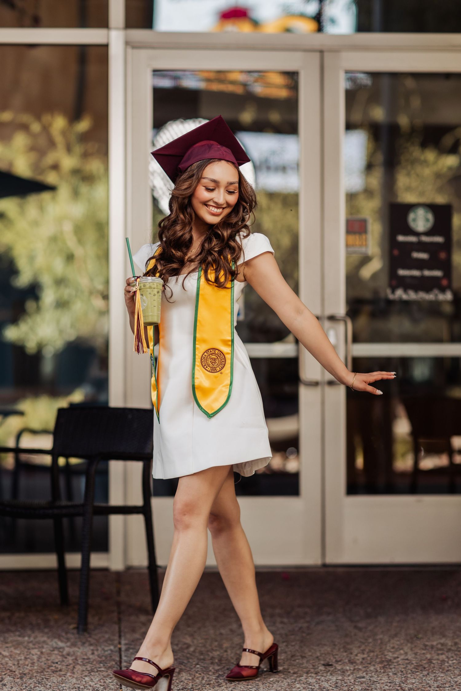 Graduate in white dress and burgundy cap poses joyfully with yellow stole and tassel during outdoor graduation photoshoot.