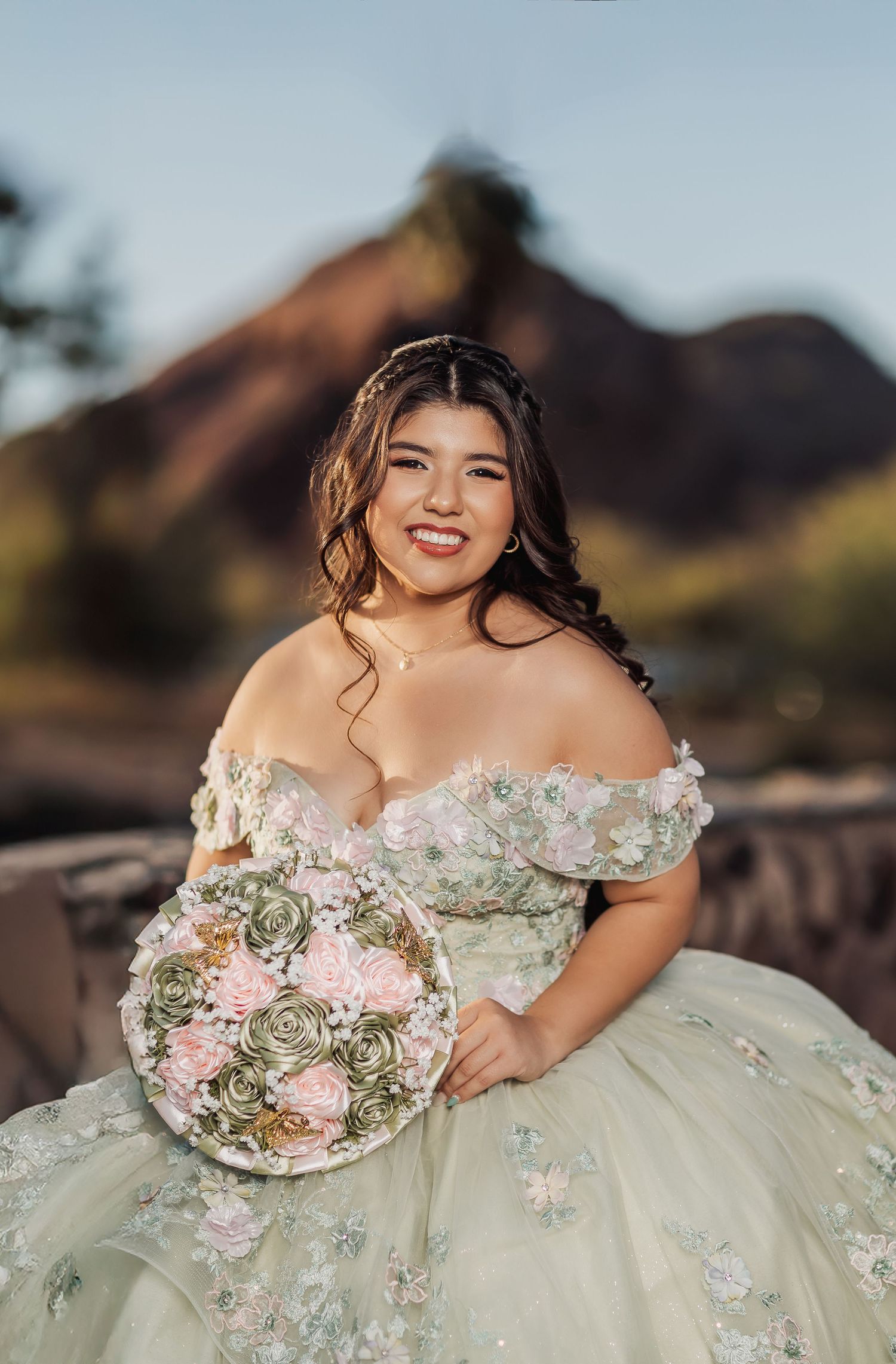 Beautiful bridal portrait in an off-shoulder floral gown holding a pink and sage bouquet against desert mountains at sunset.