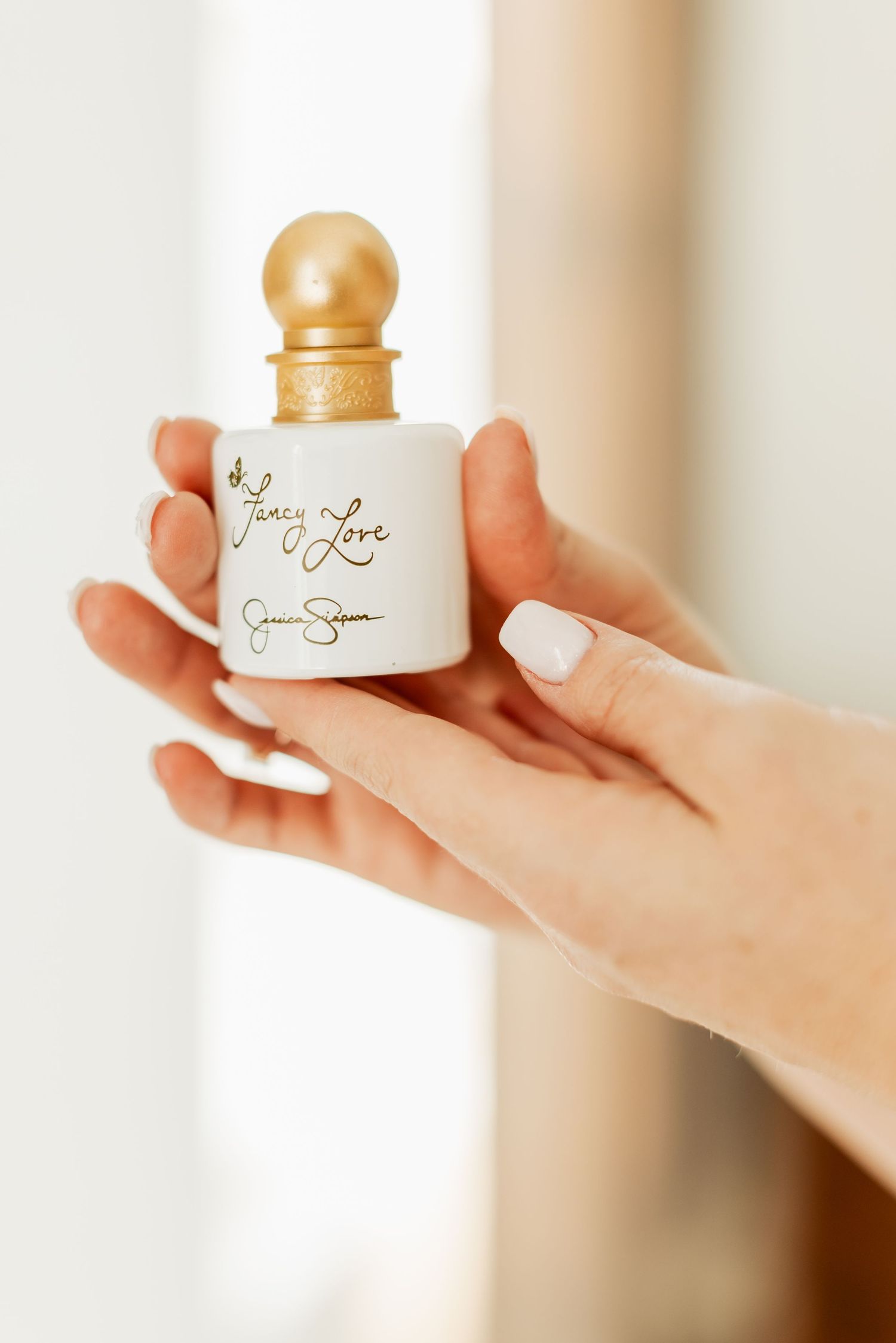 Hand holding a small white perfume bottle with gold accents against a soft, blurred background.