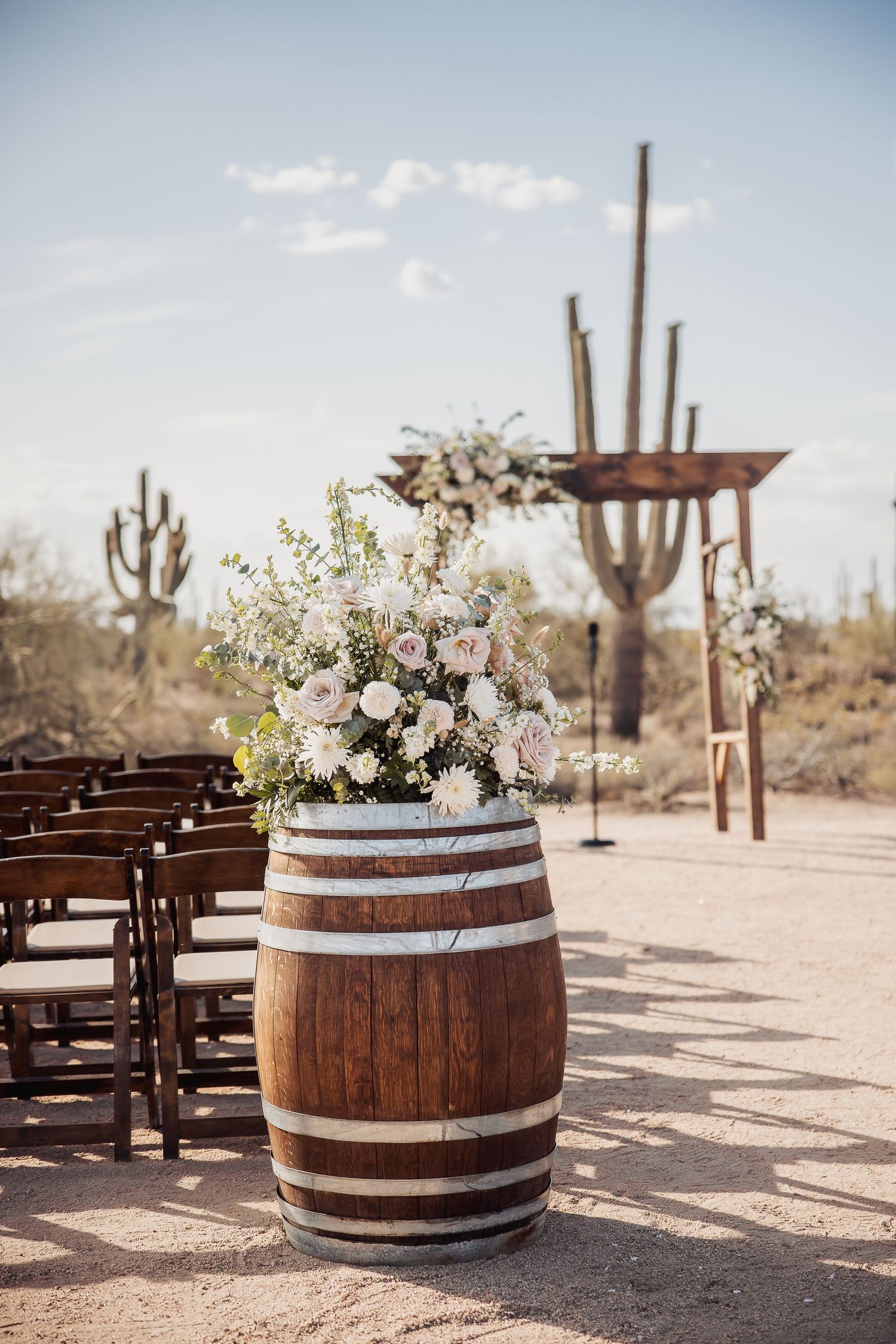 Rustic outdoor desert wedding ceremony setup with wooden barrel decor, floral arrangements and saguaro cacti in background.