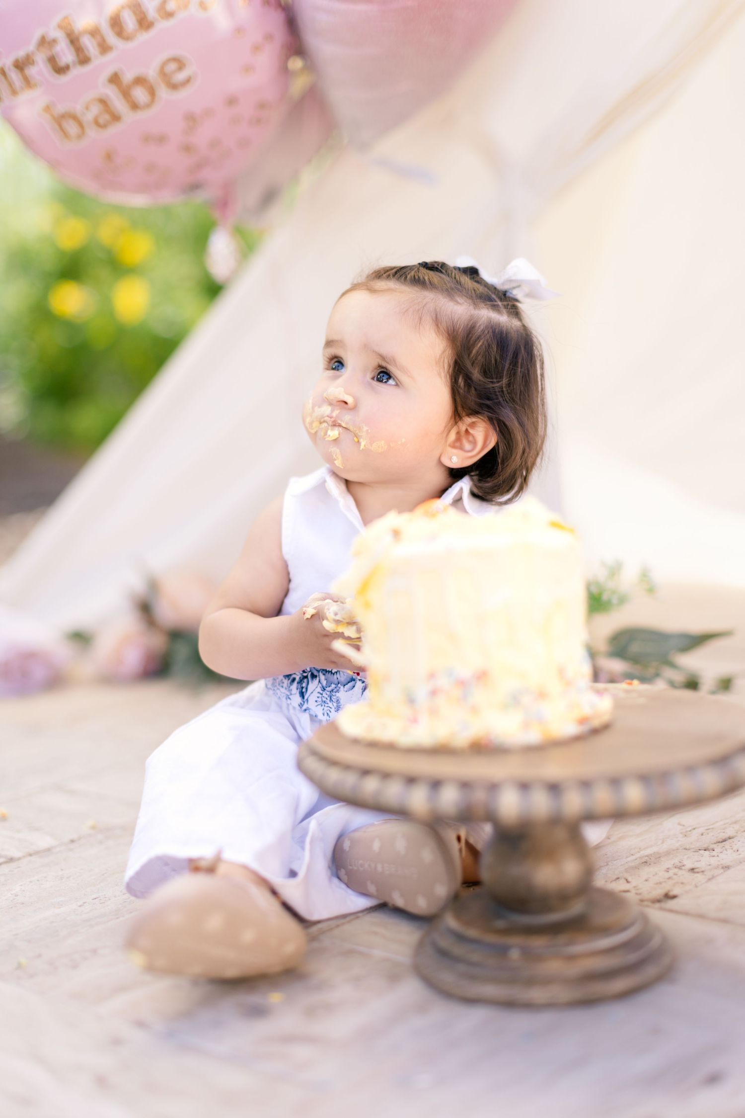 A small child wearing a white dress sits next to a yellow birthday cake on an elegant cake stand.
