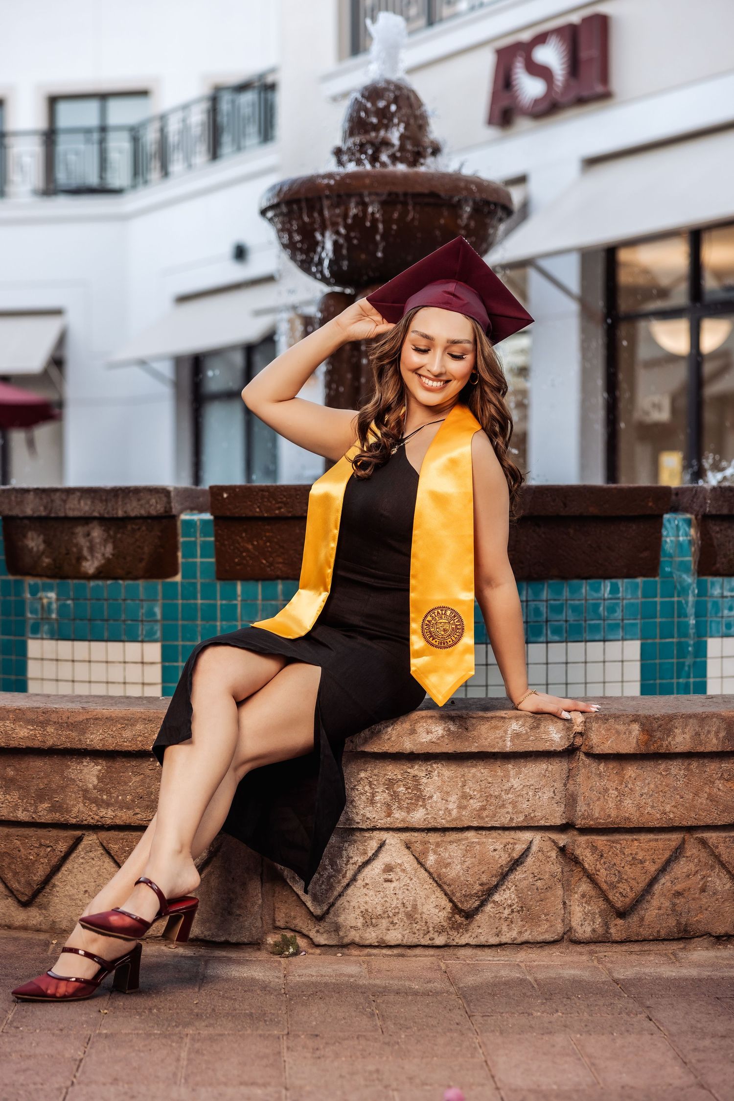 Graduate poses on stone bench wearing black dress, gold sash and maroon cap near decorative fountain.