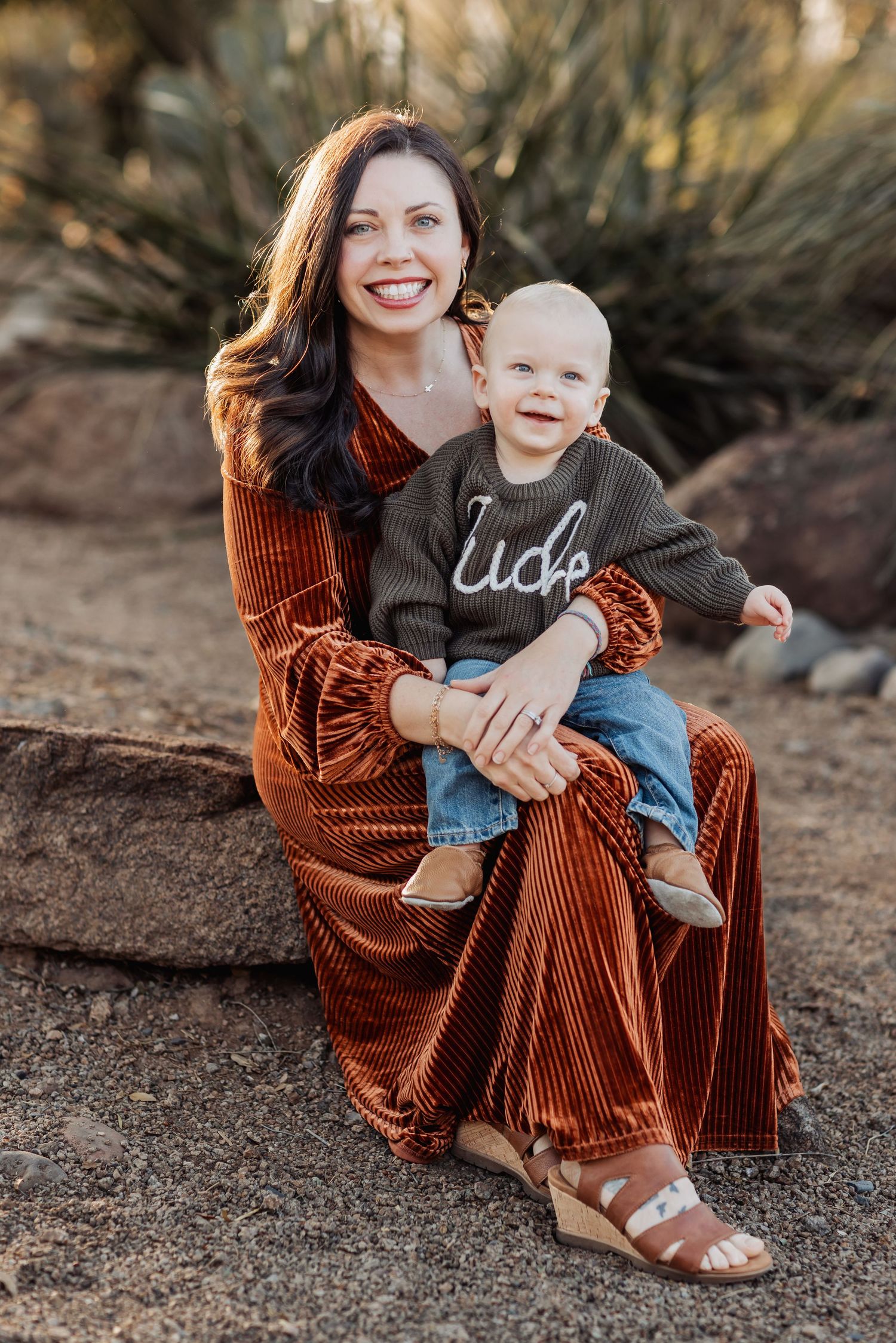A mother in a brown velvet dress hugs her small child on an outdoor path during a photo session at golden hour.