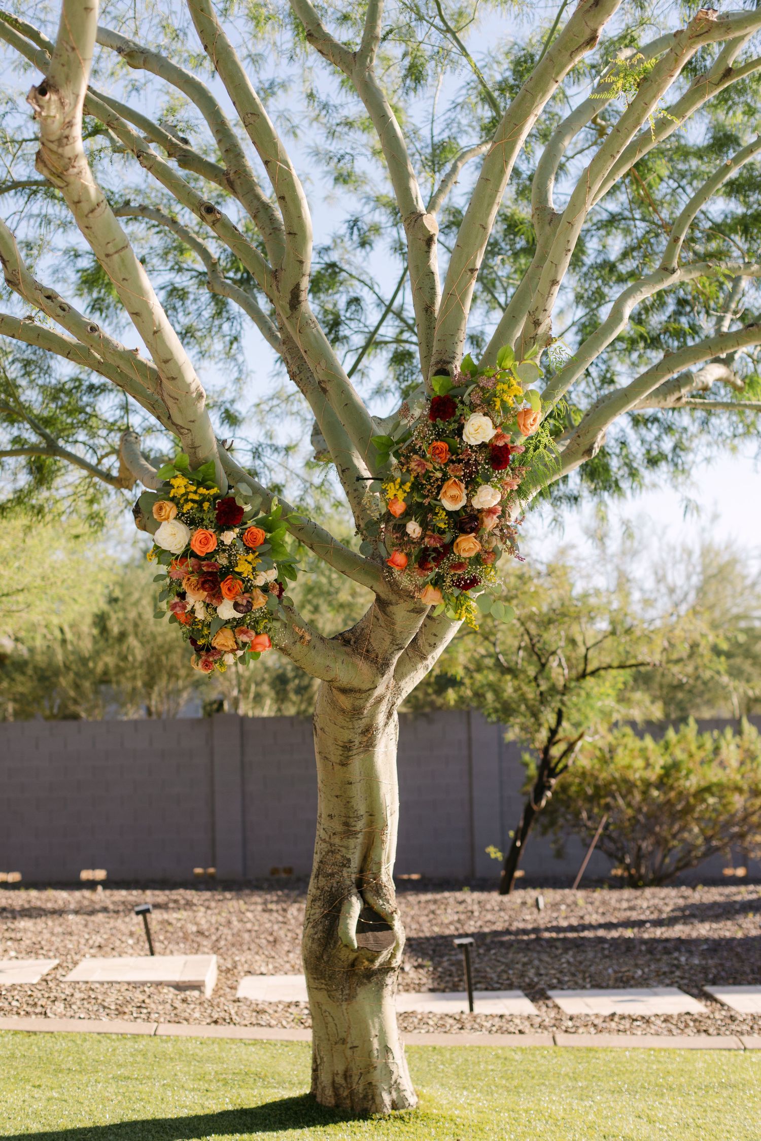 Large tree decorated with vibrant orange and pink floral arrangements in a manicured backyard with grey wall.