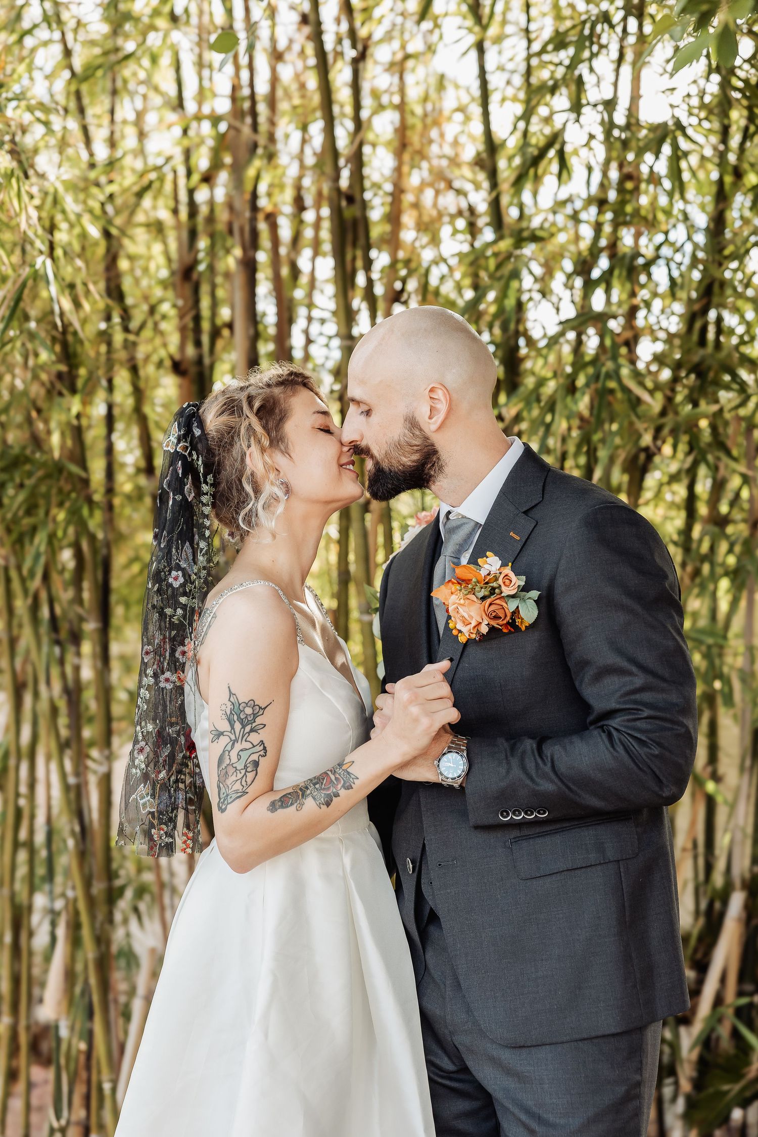A romantic wedding moment with a couple embracing in front of bamboo trees, featuring orange floral accents.
