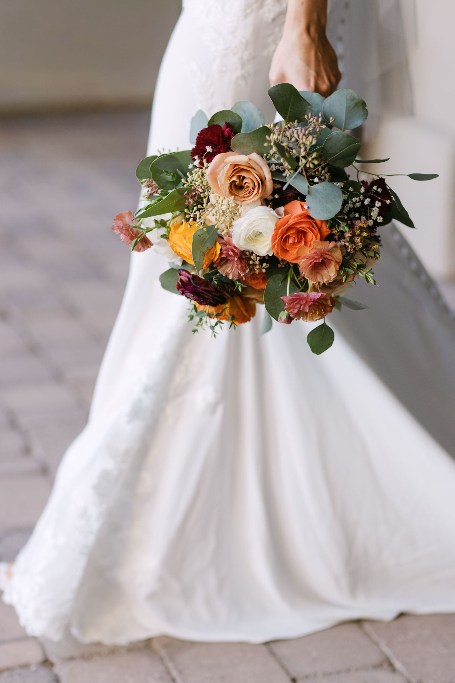 A bride in a white wedding dress holds a vibrant autumn-colored bouquet with orange and peach roses against a light backdrop.