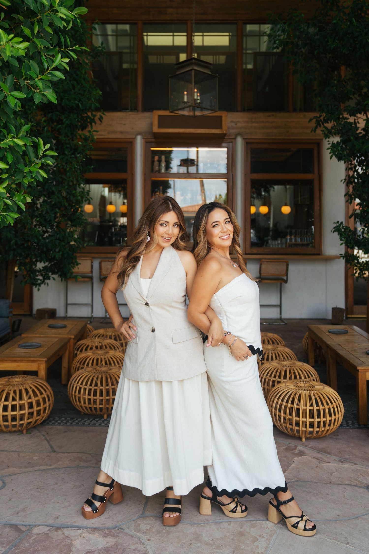 Two women in white dresses pose casually on rattan furniture against a warm wooden architectural backdrop.