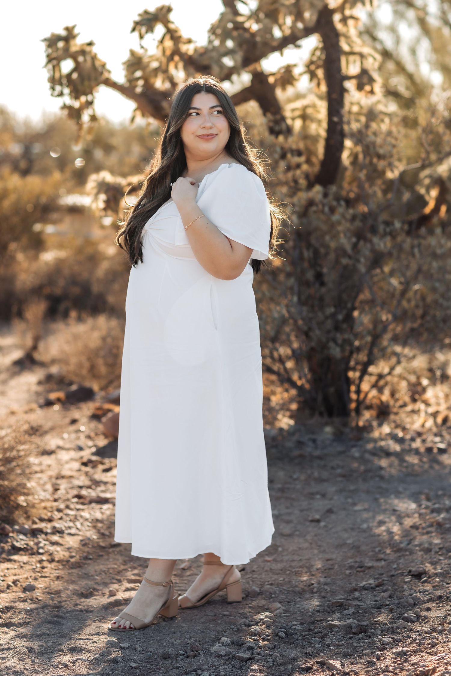 Person in flowing white dress poses outdoors in desert landscape with sun shining through trees at golden hour.