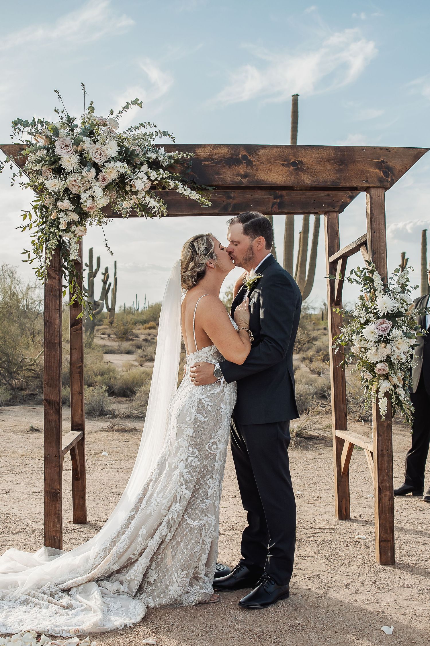 A couple shares an intimate desert wedding ceremony under a wooden pergola arch decorated with white flowers.