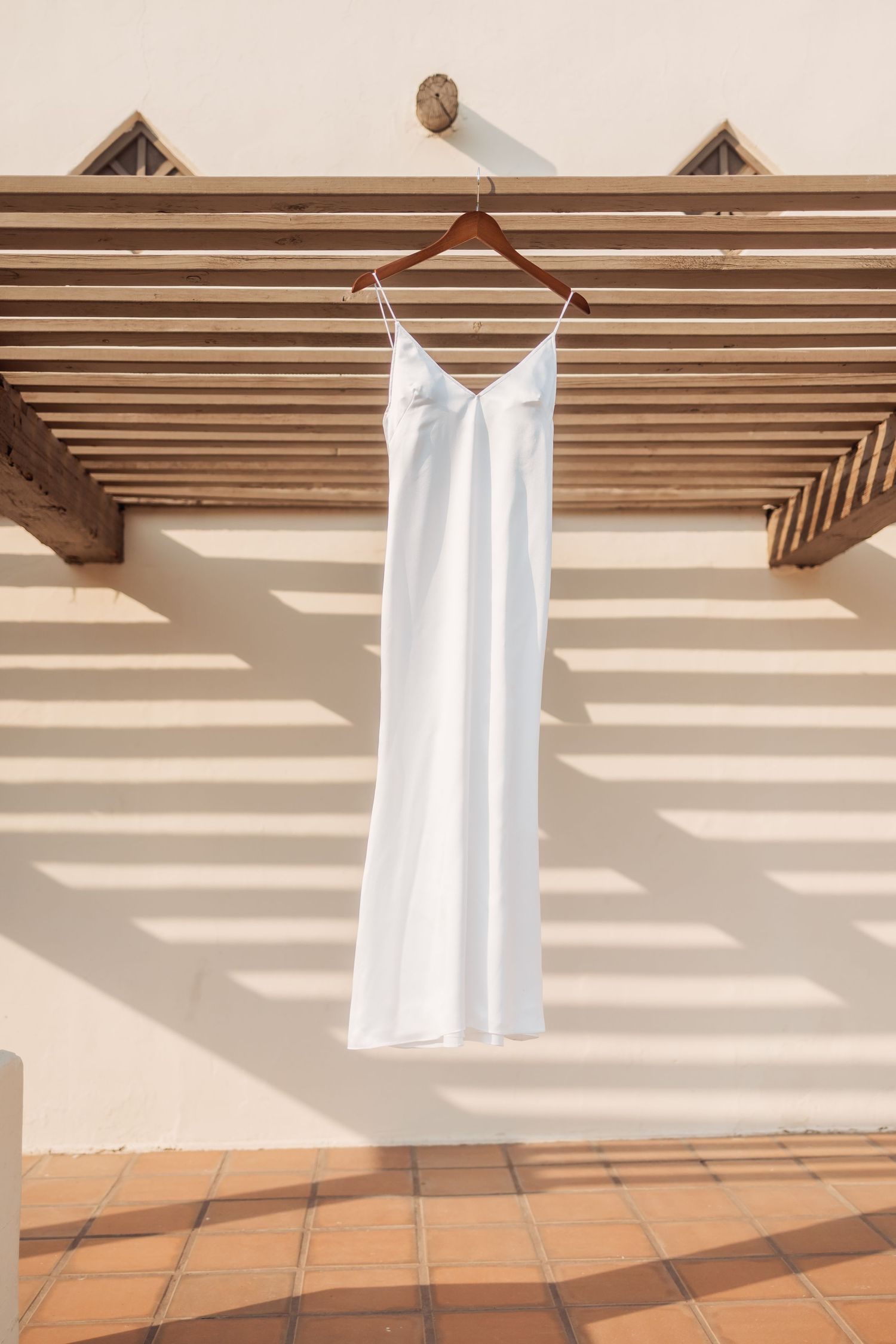 Simple white slip dress hanging on wooden hanger against a slatted wall with dramatic shadows.