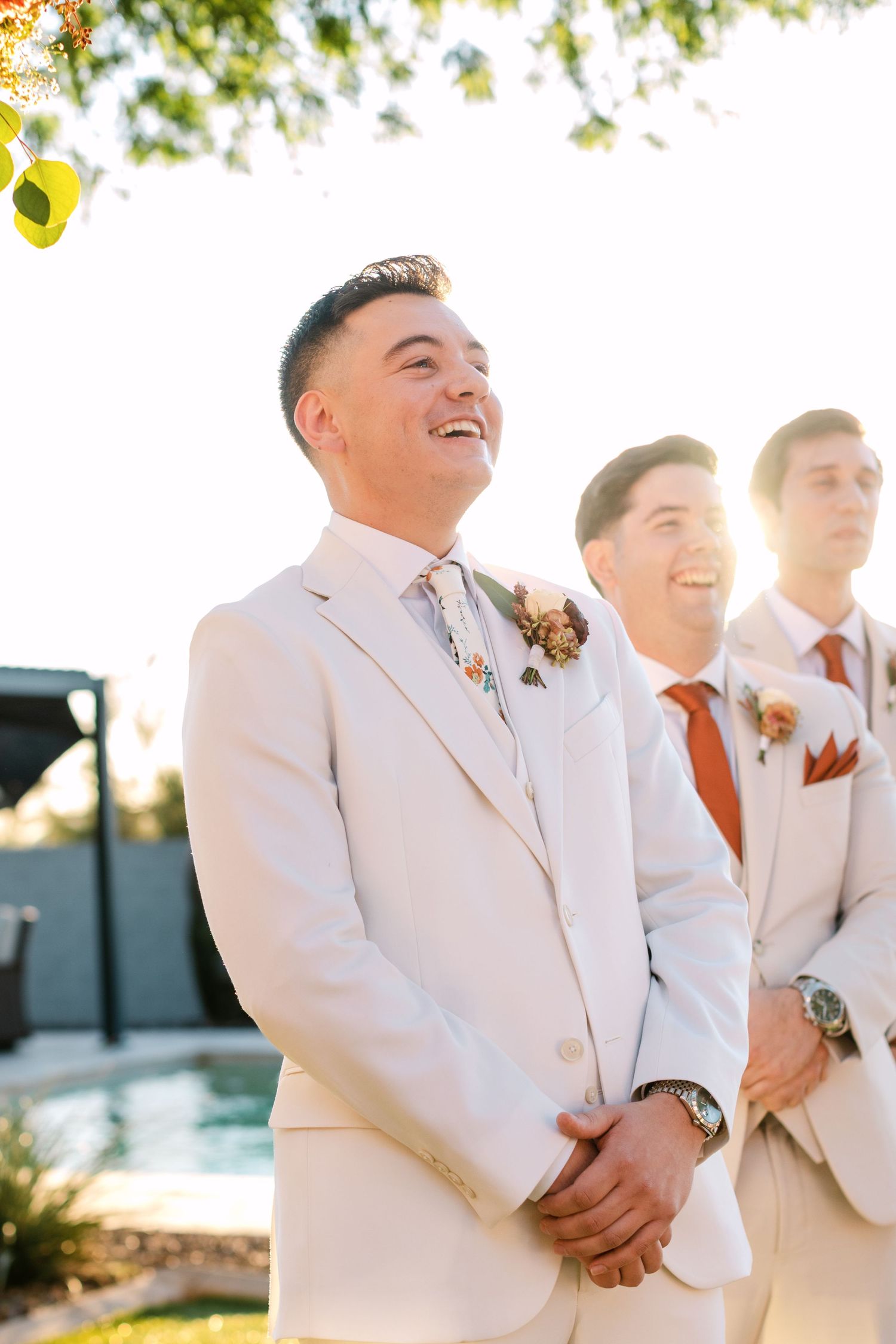 Groomsmen in white suits laughing together during outdoor wedding ceremony at sunset.