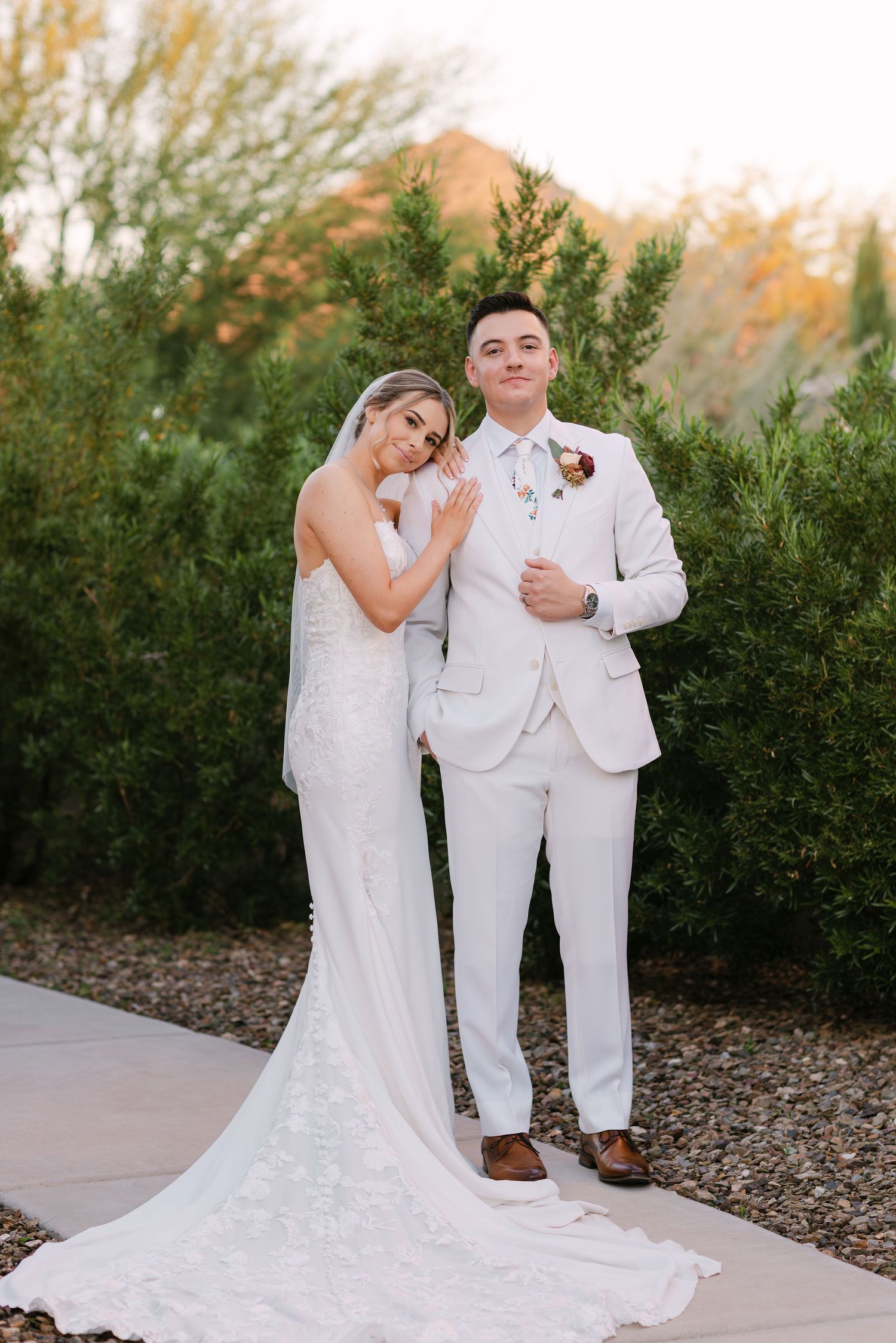 Newlywed couple in matching white outfits pose together against a backdrop of green shrubs on their wedding day.