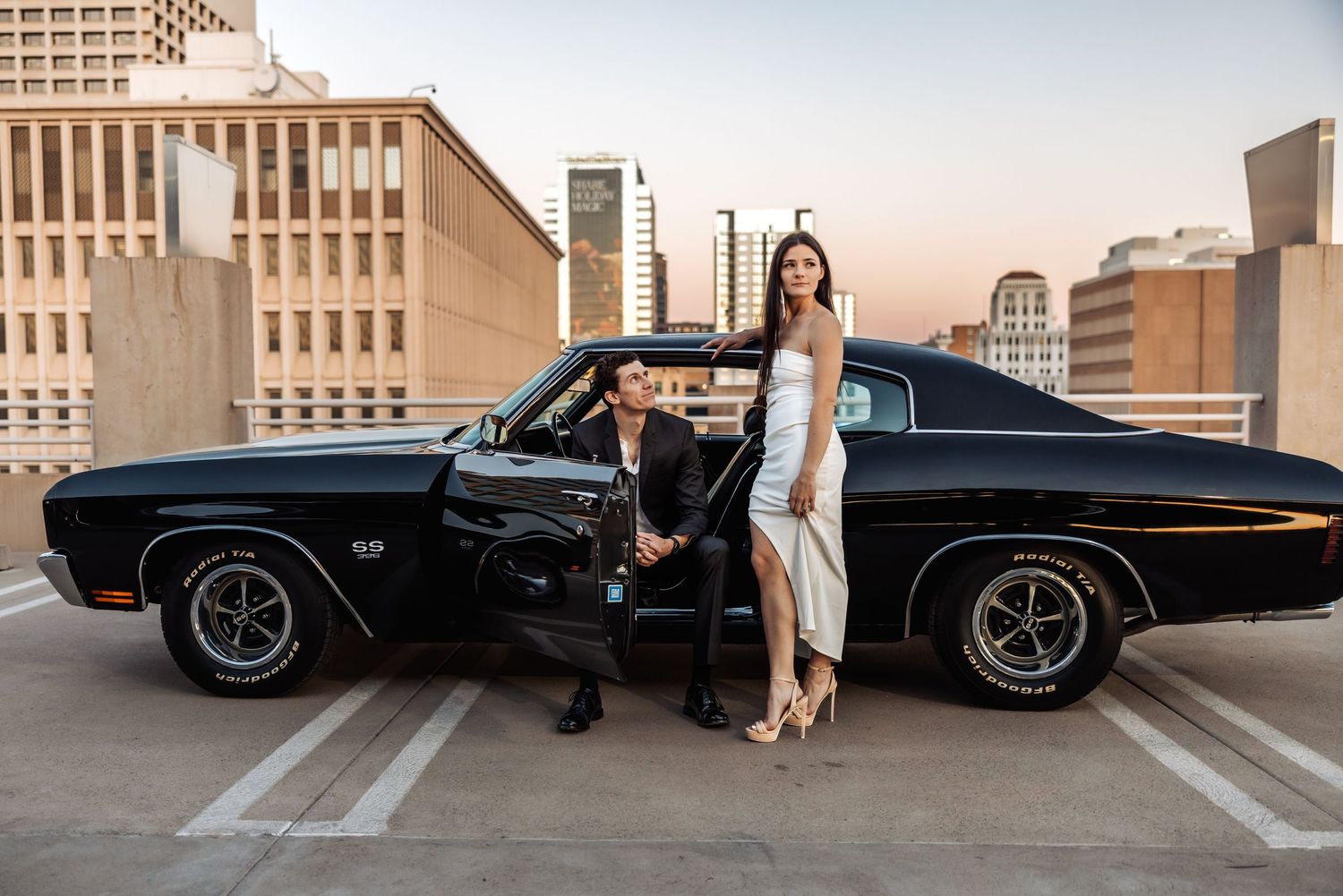 Stylish vintage black car on rooftop parking lot with city skyline backdrop during sunset.