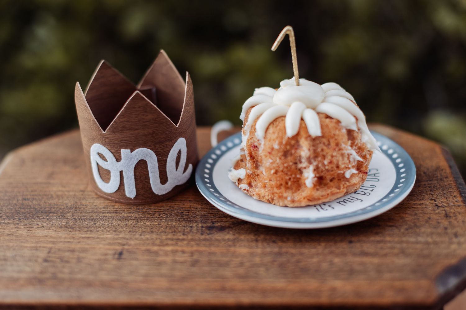 Mini bundt cake with white glaze and brown paper crown with 'one' text on wooden table.